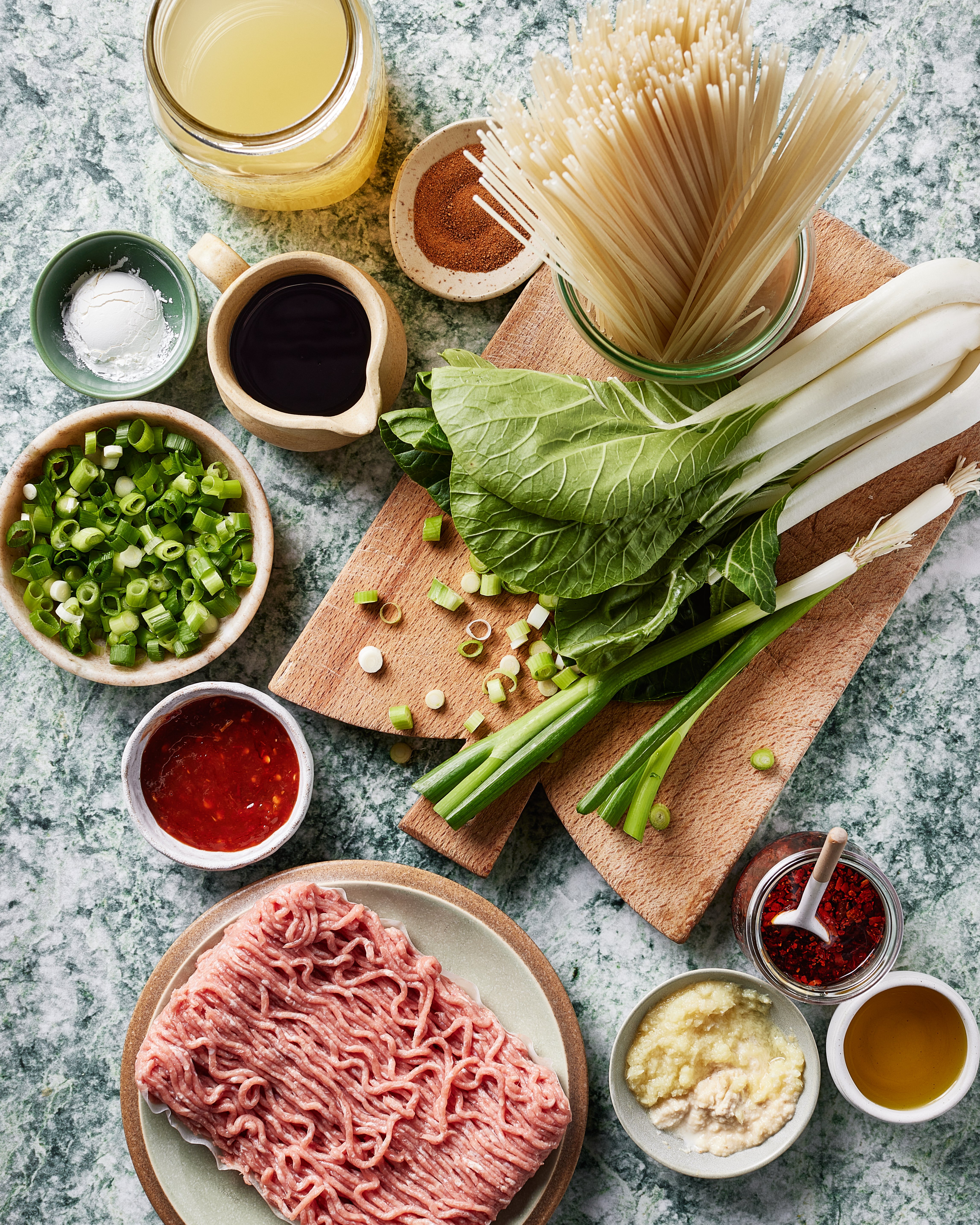 Overhead view of various ingredients on a countertop, including ground meat, bok choy, green onions, dry noodles, broth, sauces, spices, chopped garlic, and oil, all arranged neatly on dishes and boards.