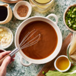 A hand uses a whisk to mix brown sauce in a white bowl, surrounded by ingredients like ground meat, broth, soy sauce, spices, minced garlic, chopped green onions, oil, noodles, and leafy greens on a marble surface.