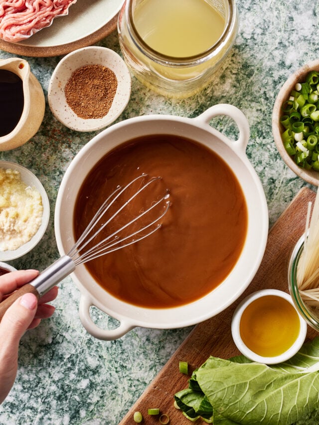 A hand uses a whisk to mix brown sauce in a white bowl, surrounded by ingredients like ground meat, broth, soy sauce, spices, minced garlic, chopped green onions, oil, noodles, and leafy greens on a marble surface.