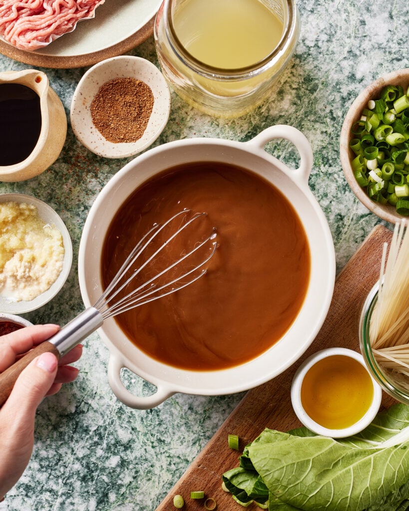 A hand uses a whisk to mix brown sauce in a white bowl, surrounded by ingredients like ground meat, broth, soy sauce, spices, minced garlic, chopped green onions, oil, noodles, and leafy greens on a marble surface.
