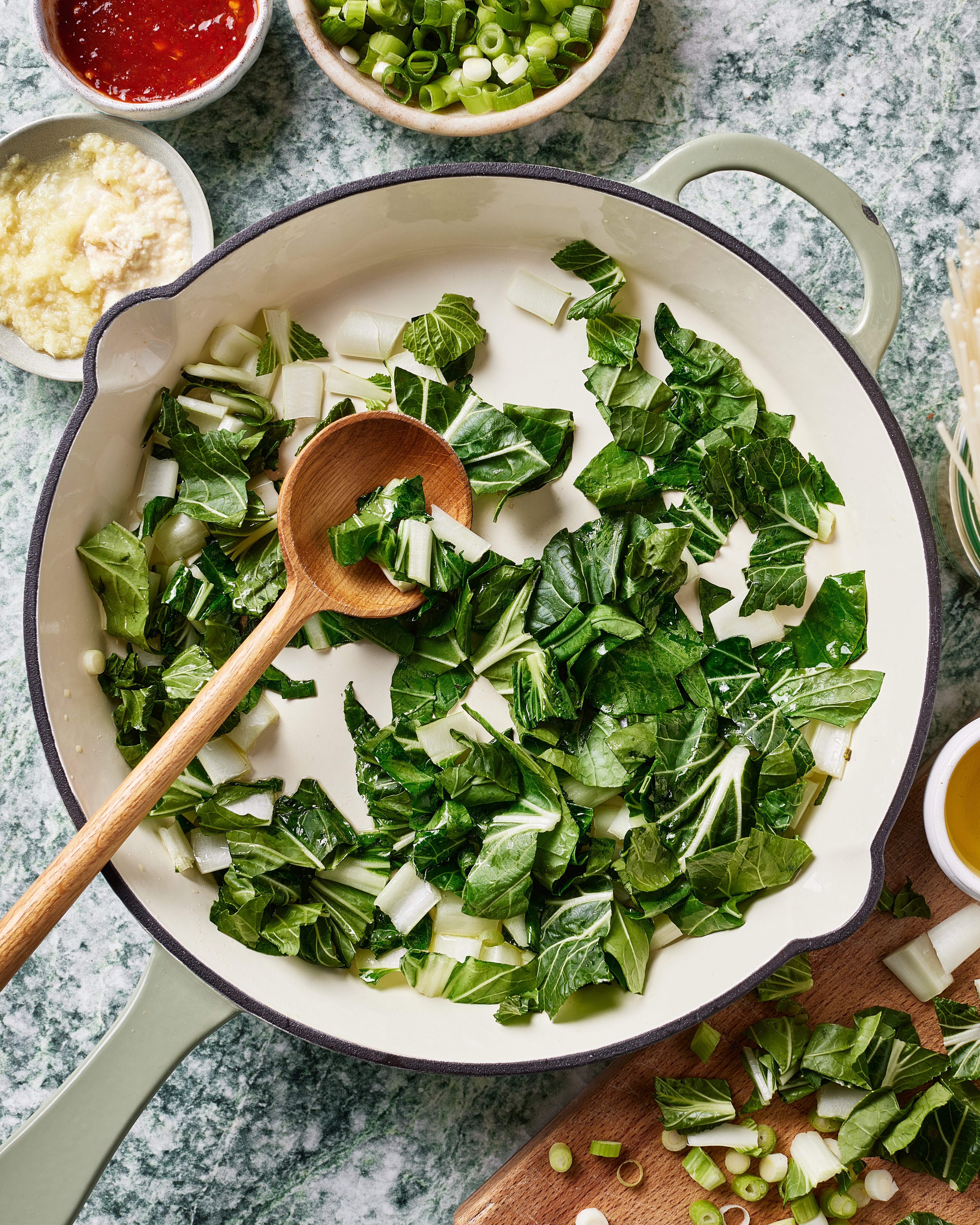 A large skillet with chopped bok choy being sautéed, stirred with a wooden spoon. Surrounding the skillet are bowls of chopped green onions, minced garlic, a red sauce, and a cutting board with more vegetables.