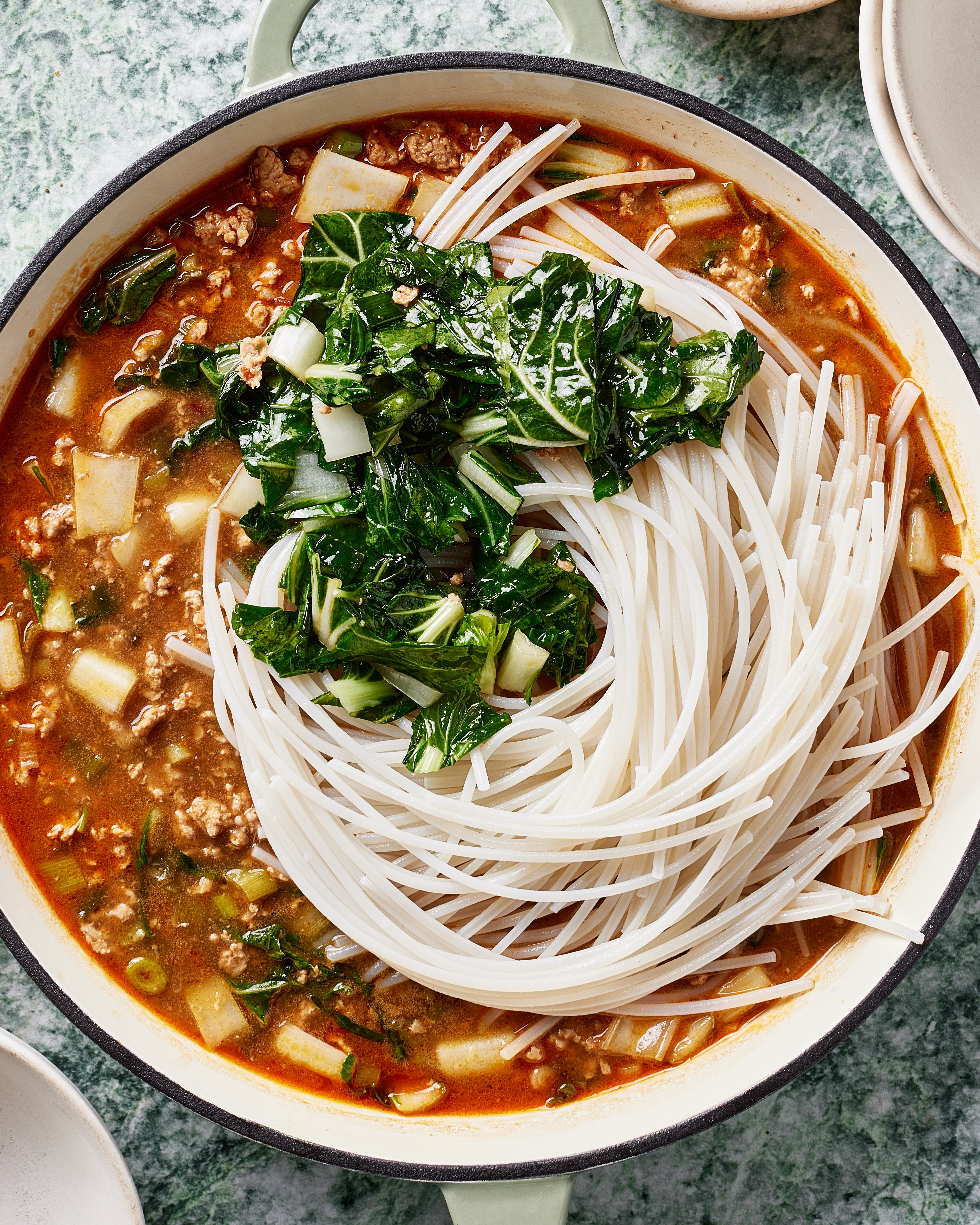 A pot of noodle soup with uncooked rice noodles, leafy greens, minced meat, and chopped vegetables in a rich, reddish broth, placed on a green textured surface.