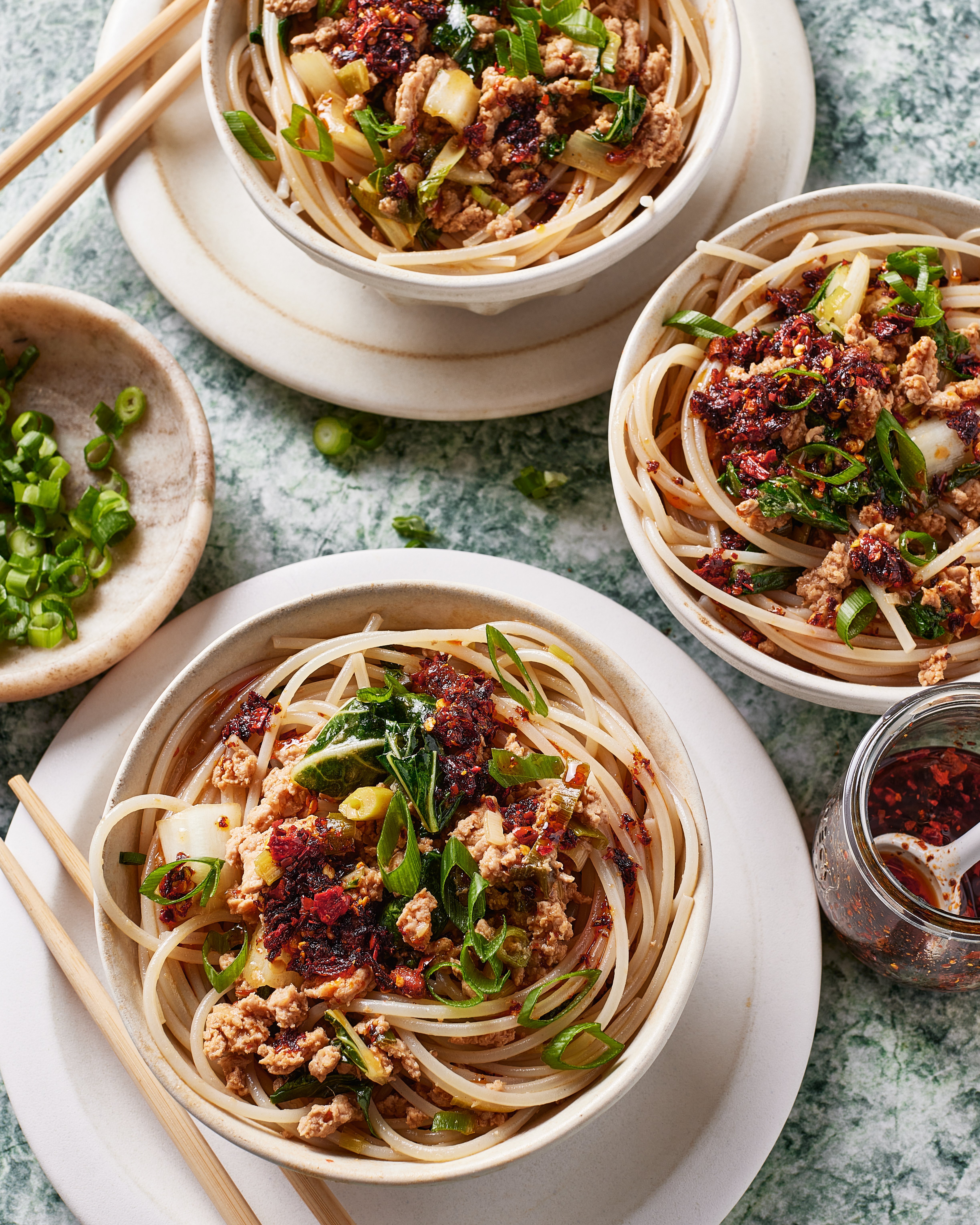 Three bowls of noodles topped with minced meat, chopped green onions, and chili oil are arranged on a table with chopsticks and a small dish of sliced green onions nearby.