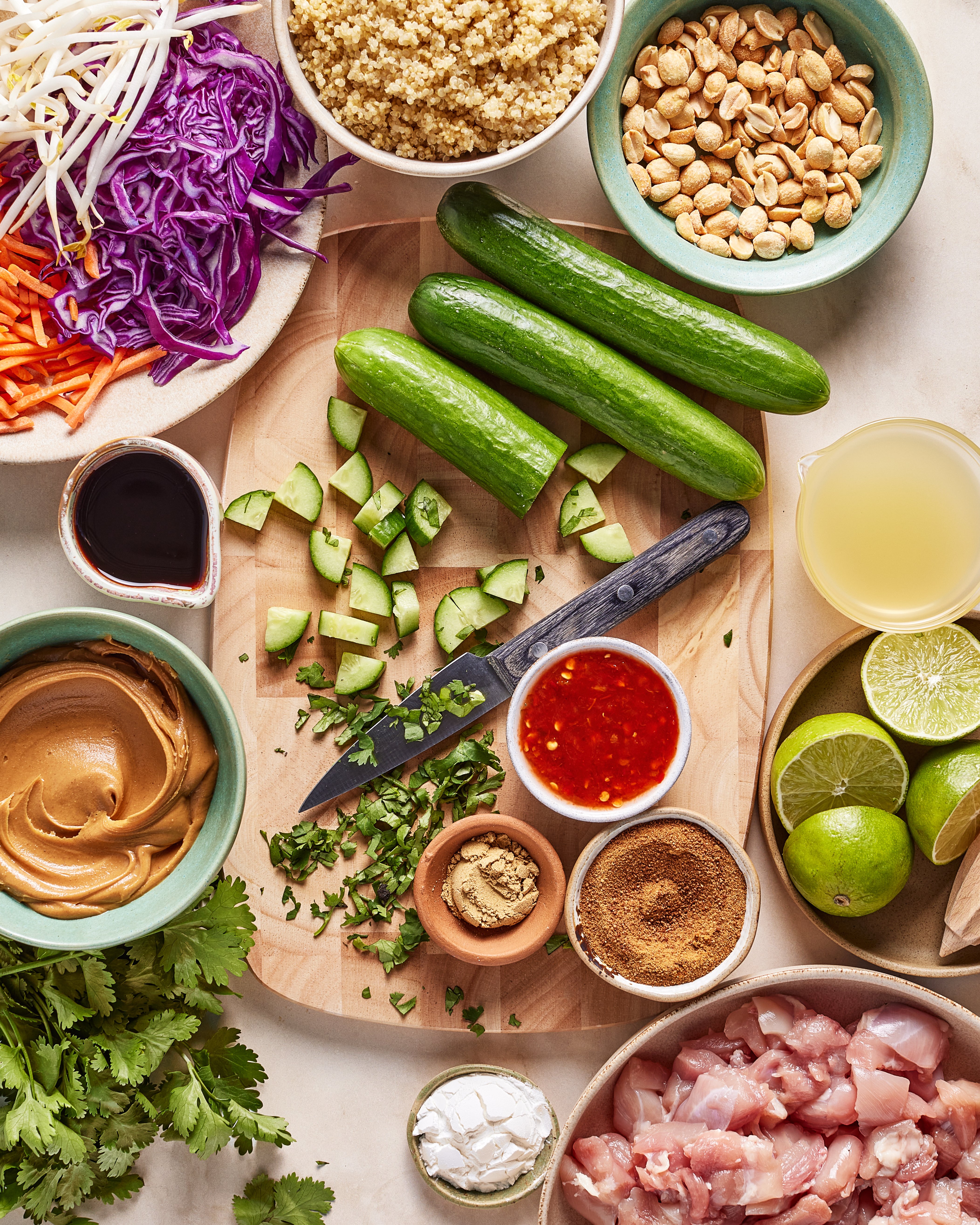 A top-down view of fresh ingredients on a counter, including cucumbers, limes, purple cabbage, peanuts, chicken, quinoa, cilantro, peanut butter, sauces, and spices, with a knife and chopped cucumber on a cutting board.
