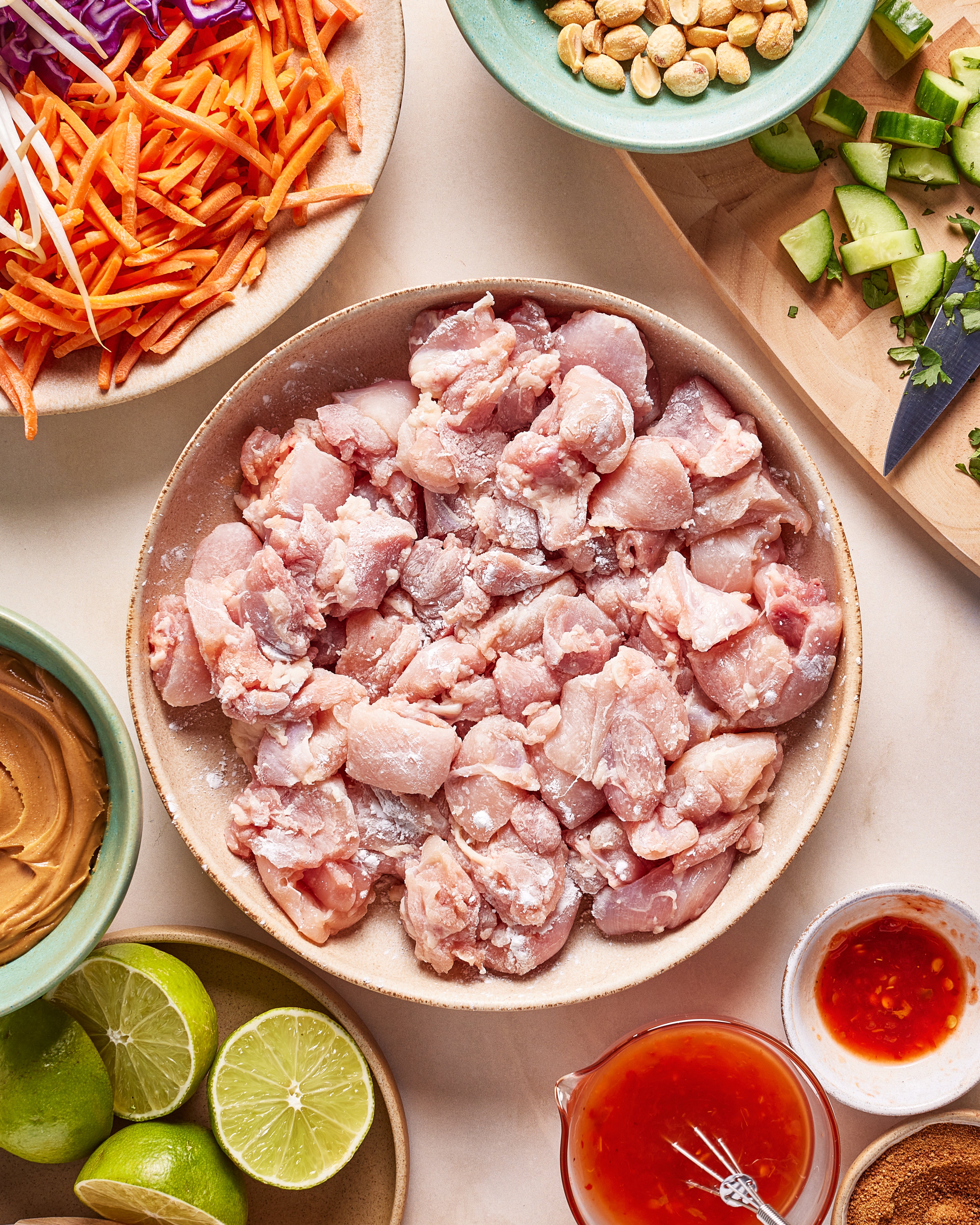 A bowl of raw, diced chicken surrounded by sliced carrots, cucumber, peanuts, lime halves, peanut butter, red sauce, and shredded purple cabbage on a table.