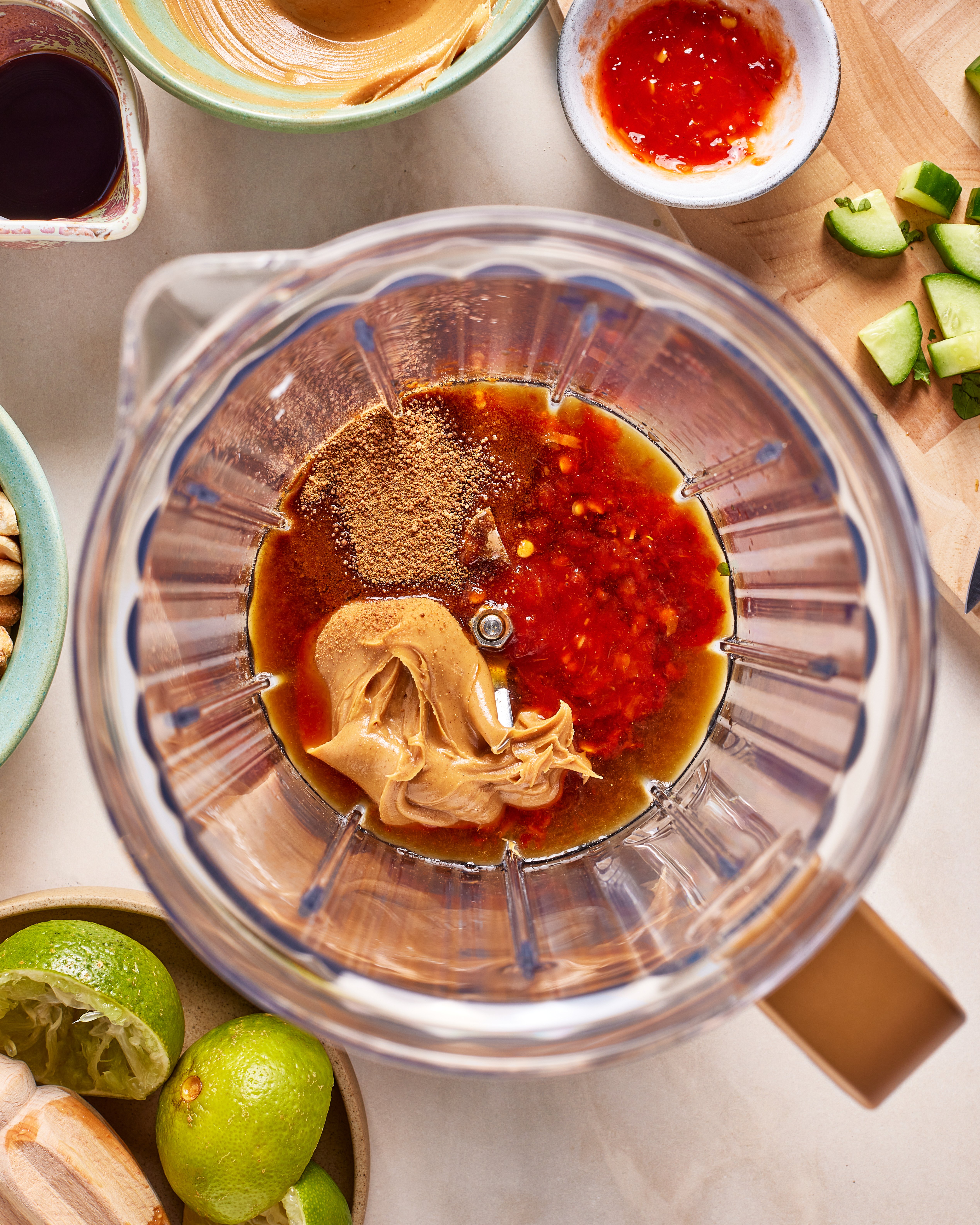 A blender filled with peanut butter, soy sauce, chili sauce, and spices sits on a countertop surrounded by bowls of limes, chopped avocado, peanut butter, and chili sauce.
