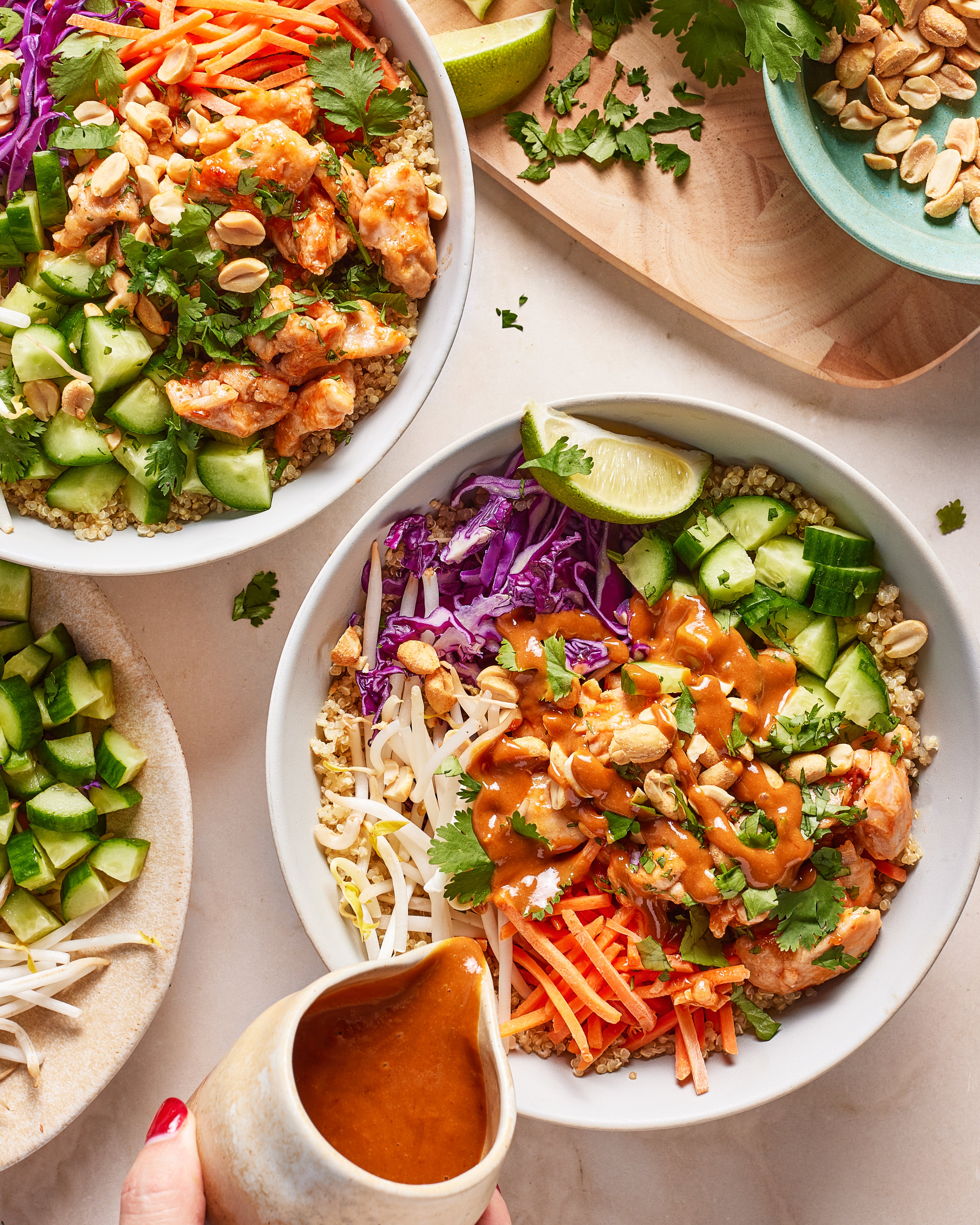 A hand pours peanut sauce onto a bowl filled with colorful veggies, including carrots, cabbage, cucumber, cilantro, and peanuts. Two similar veggie bowls and a plate of peanuts are nearby on a light surface.