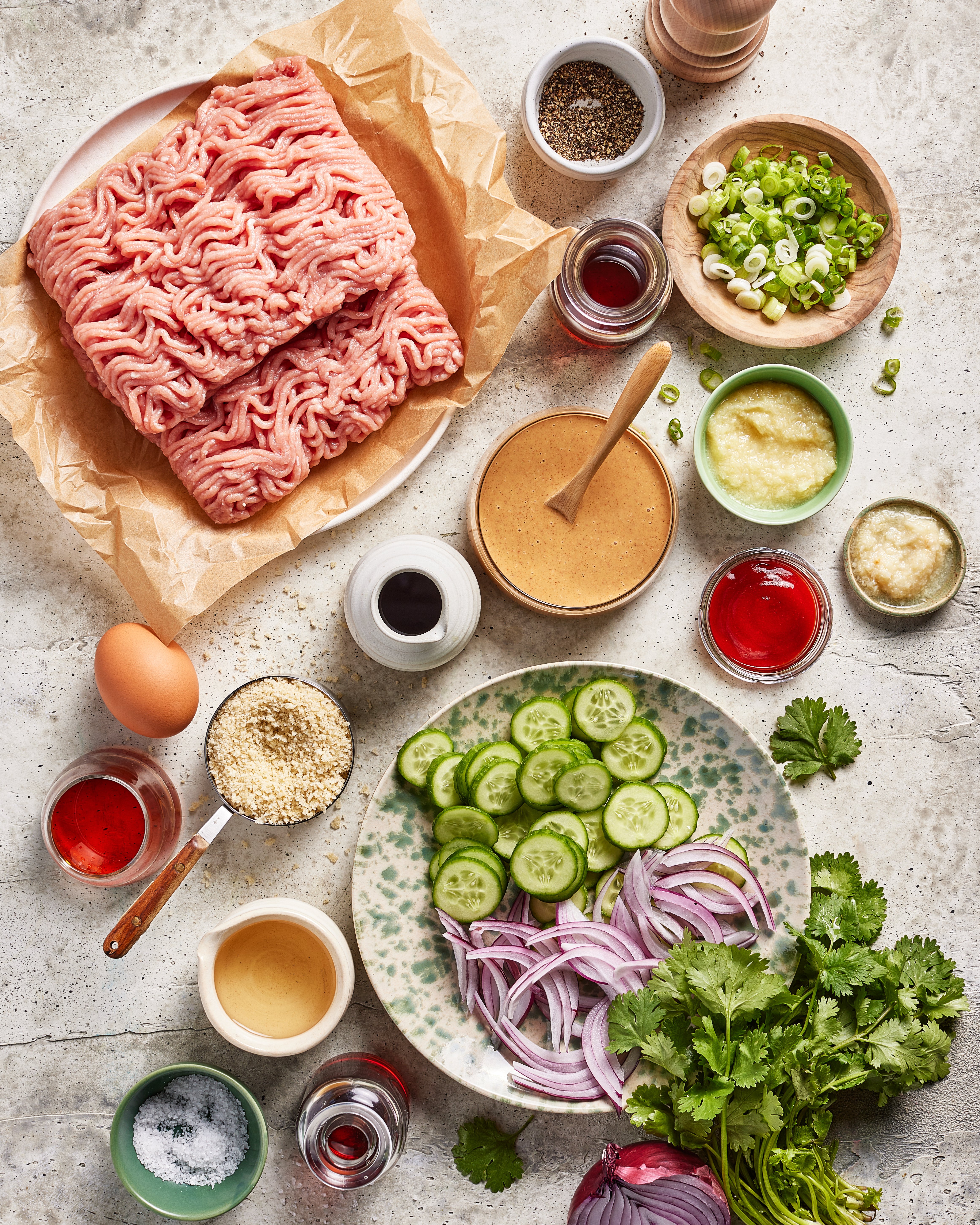 A flat lay of ingredients on a light surface, including ground meat, sliced cucumbers, red onion, cilantro, scallions, egg, sauces, seasonings, and various liquids in small bowls.