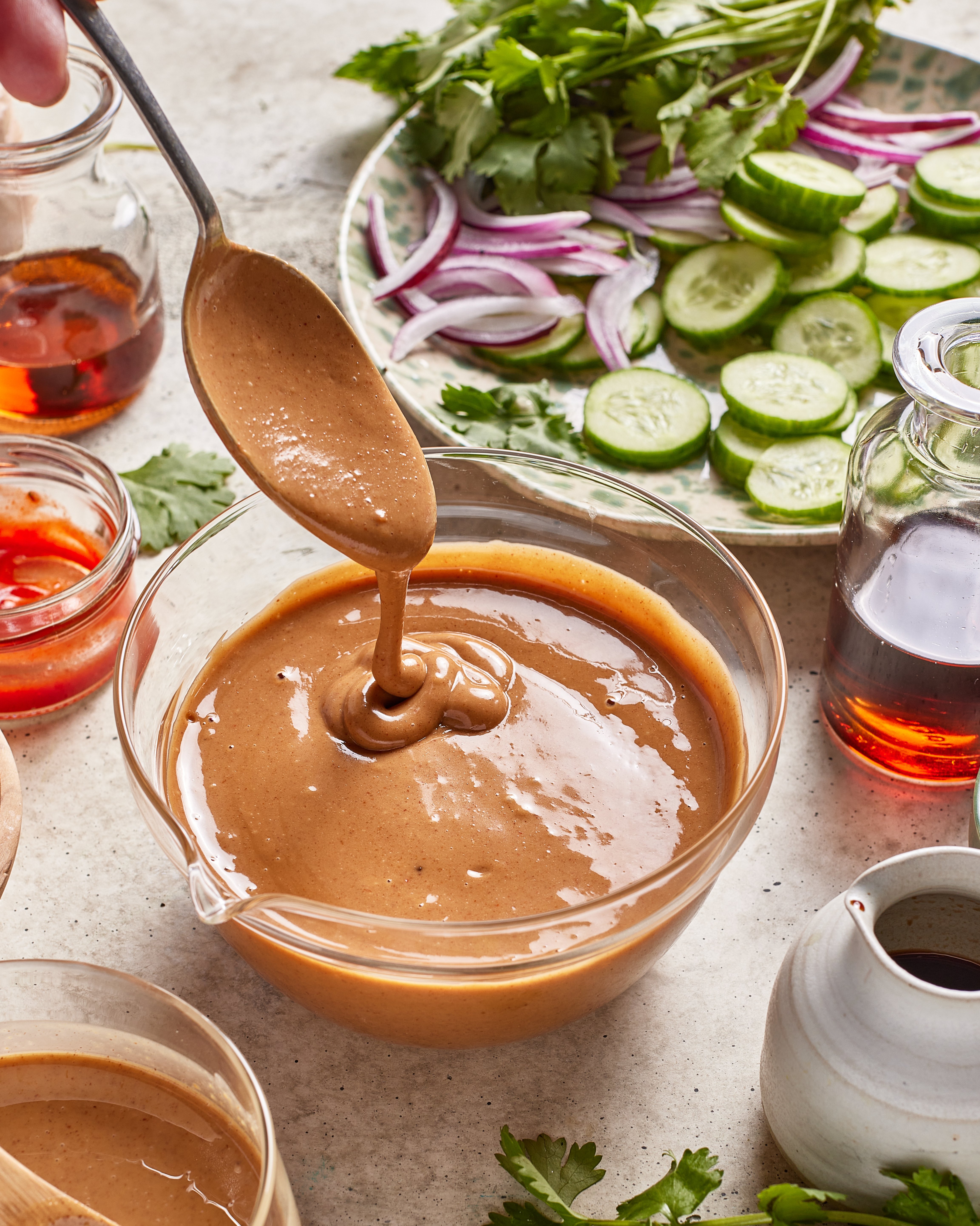 A spoon drizzles creamy brown peanut sauce into a glass bowl, surrounded by sliced cucumbers, red onions, cilantro, and jars of various liquids on a light surface.