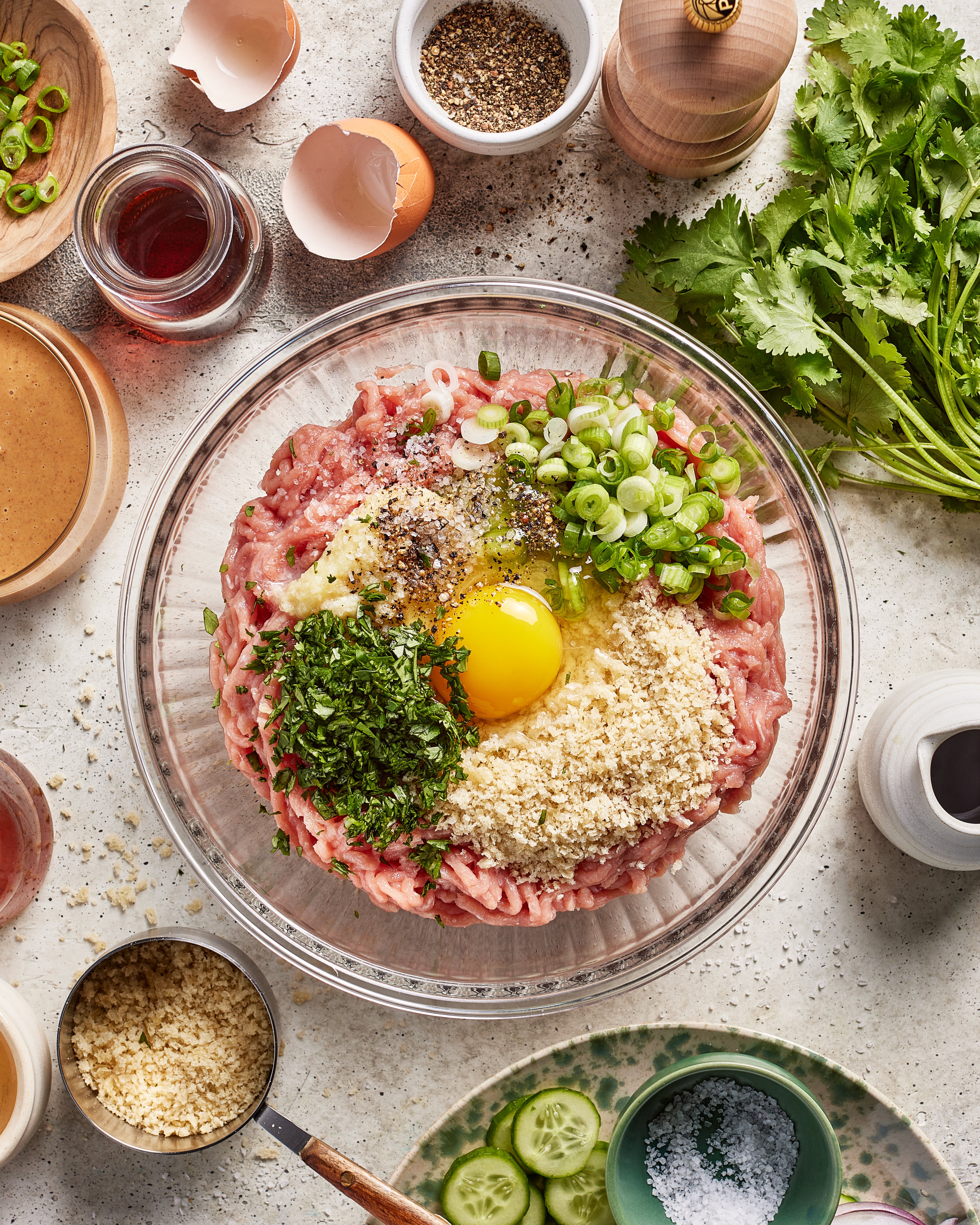 A glass bowl with ground meat, a raw egg, breadcrumbs, chopped herbs, and green onions, surrounded by ingredients like spices, cracked eggshells, cucumber slices, fresh cilantro, and seasoning bowls on a kitchen counter.