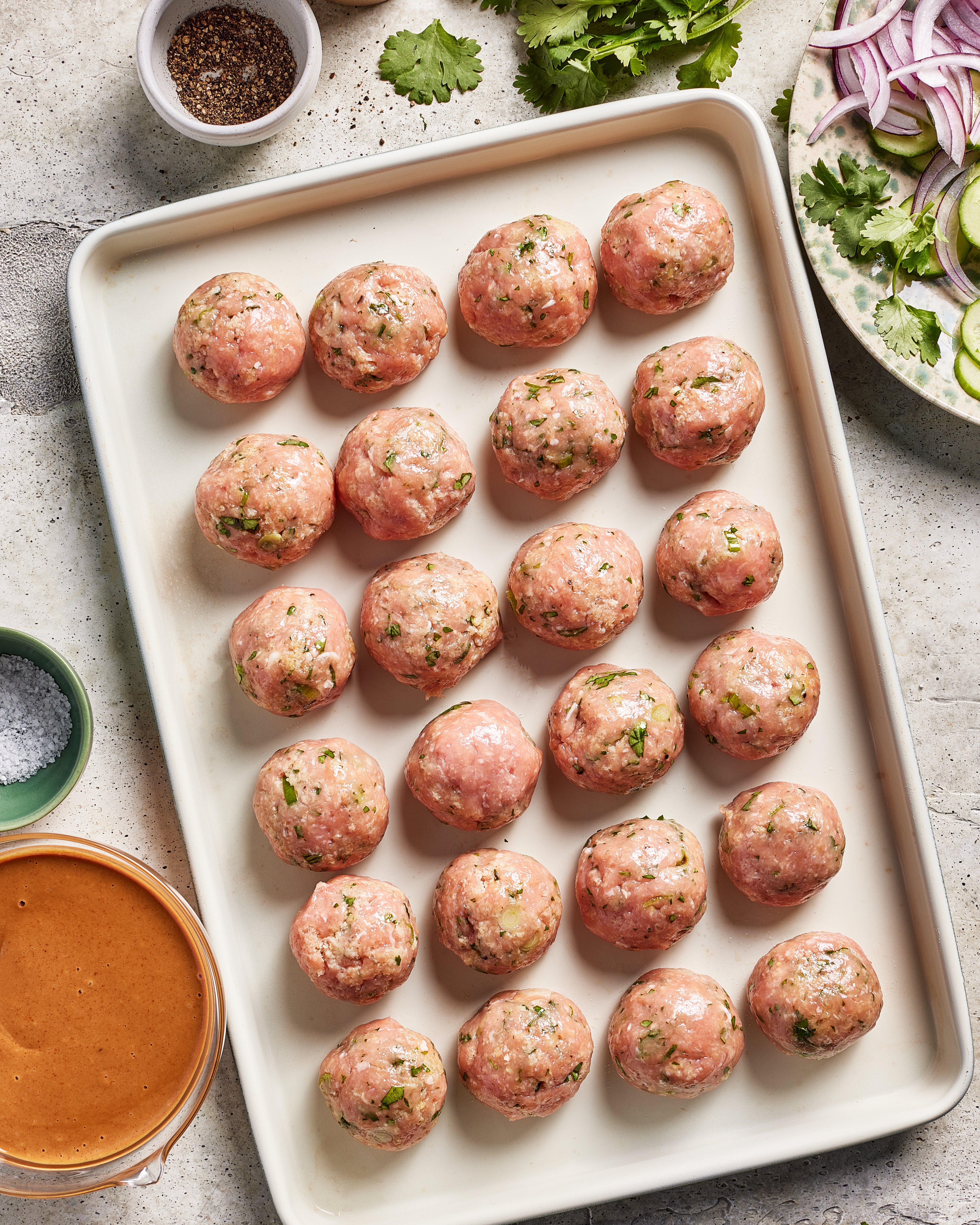 A tray of uncooked chicken dumpling meatballs mixed with herbs is arranged in rows on a white tray. Surrounding the tray are bowls of pepper, salt, a reddish sauce, and sliced red onions with cilantro.