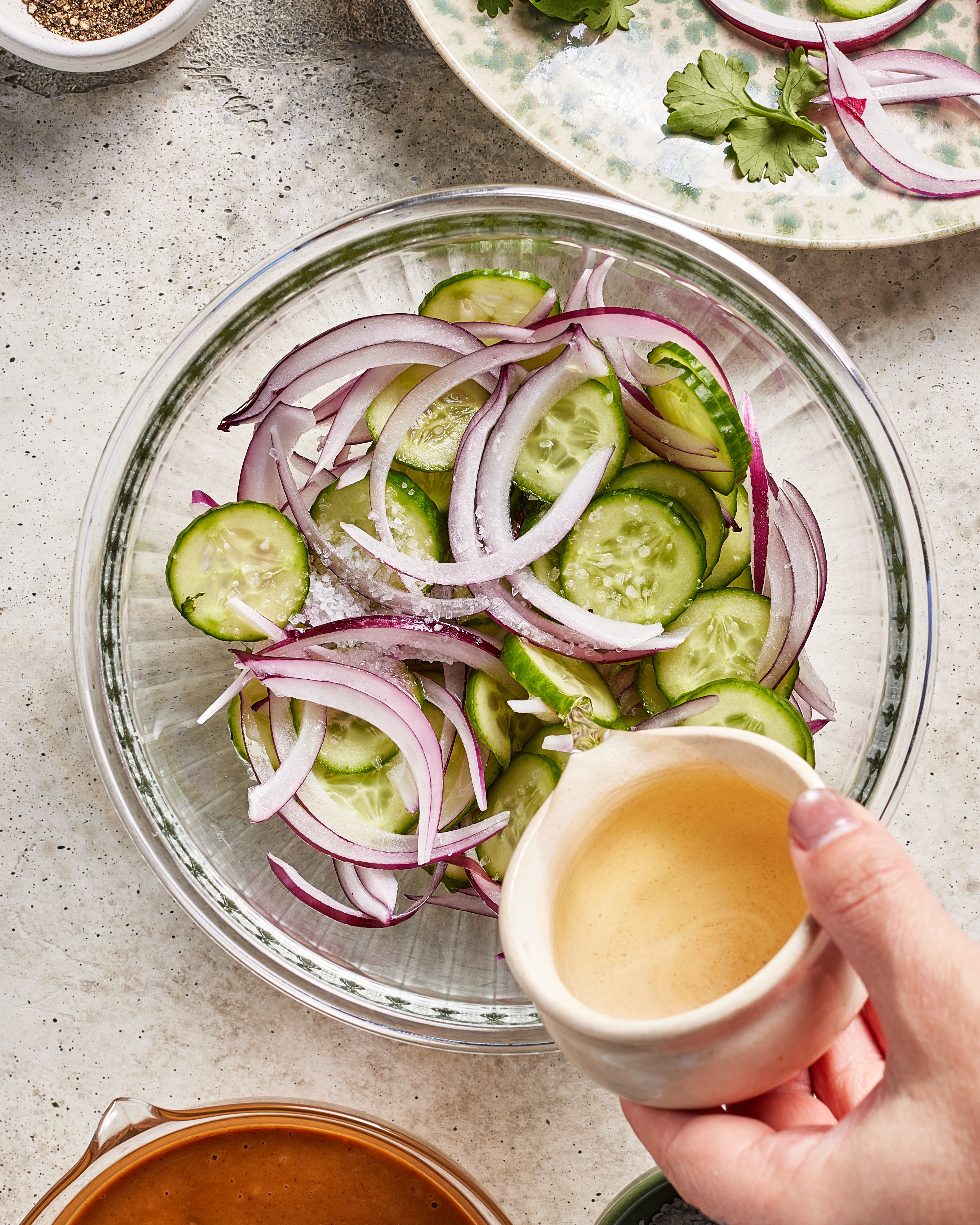 A hand pours vinegar from a small white pitcher over a glass bowl filled with sliced cucumbers and red onions, preparing a fresh salad on a light-colored countertop.