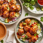 Two bowls of rice topped with chicken dumpling meatballs, sliced cucumbers, red onion, and creamy sauce, garnished with chili flakes. Fresh cilantro, cucumber slices, and a bowl of sauce are arranged nearby.
