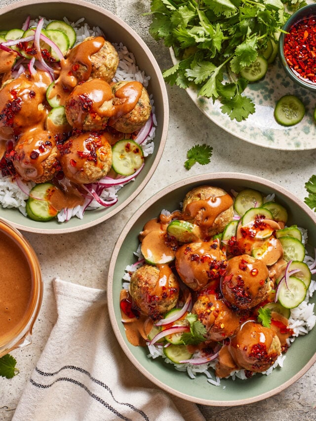 Two bowls of rice topped with chicken dumpling meatballs, sliced cucumbers, red onion, and creamy sauce, garnished with chili flakes. Fresh cilantro, cucumber slices, and a bowl of sauce are arranged nearby.
