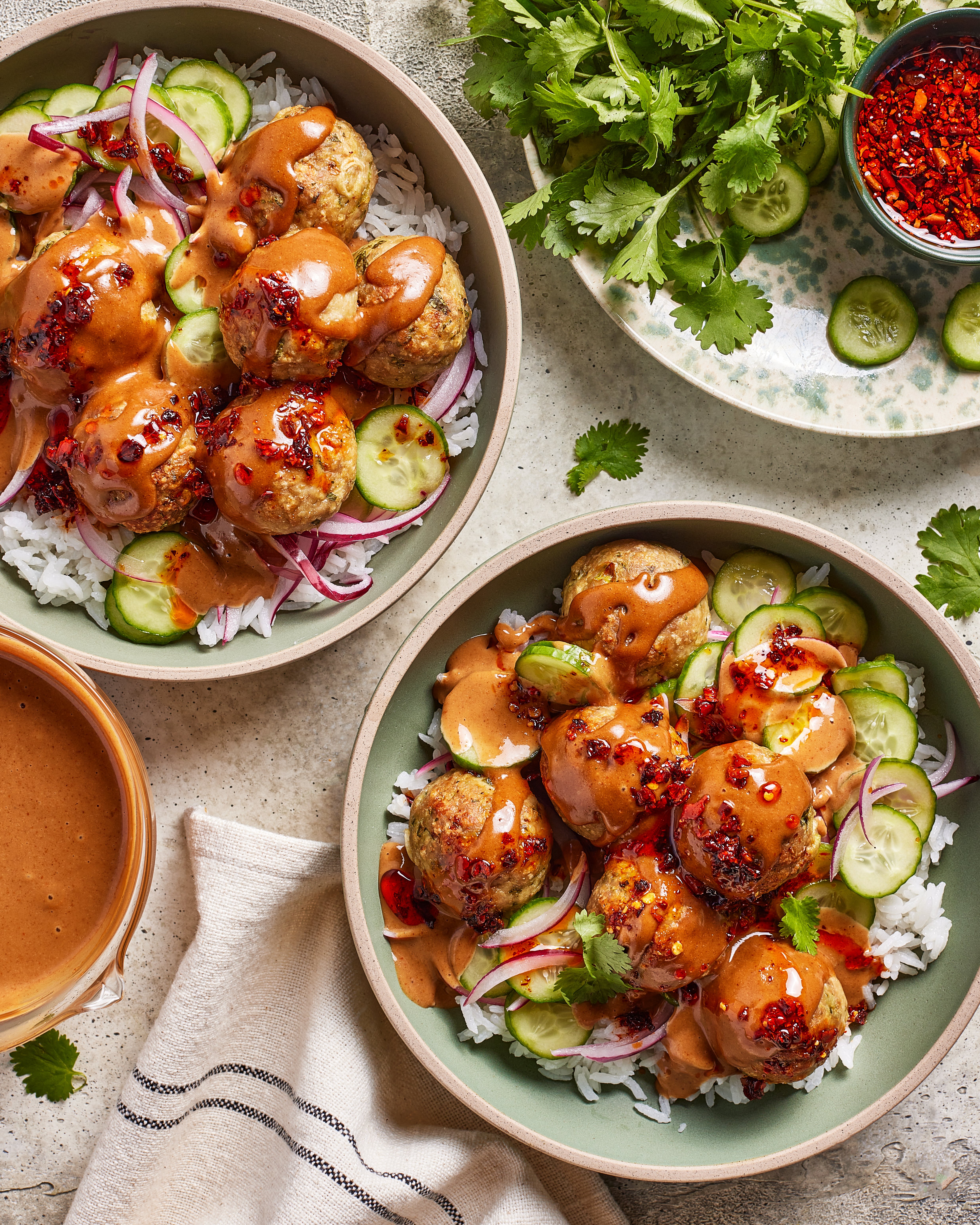 Two bowls of rice topped with chicken dumpling meatballs, sliced cucumbers, red onion, and creamy sauce, garnished with chili flakes. Fresh cilantro, cucumber slices, and a bowl of sauce are arranged nearby.