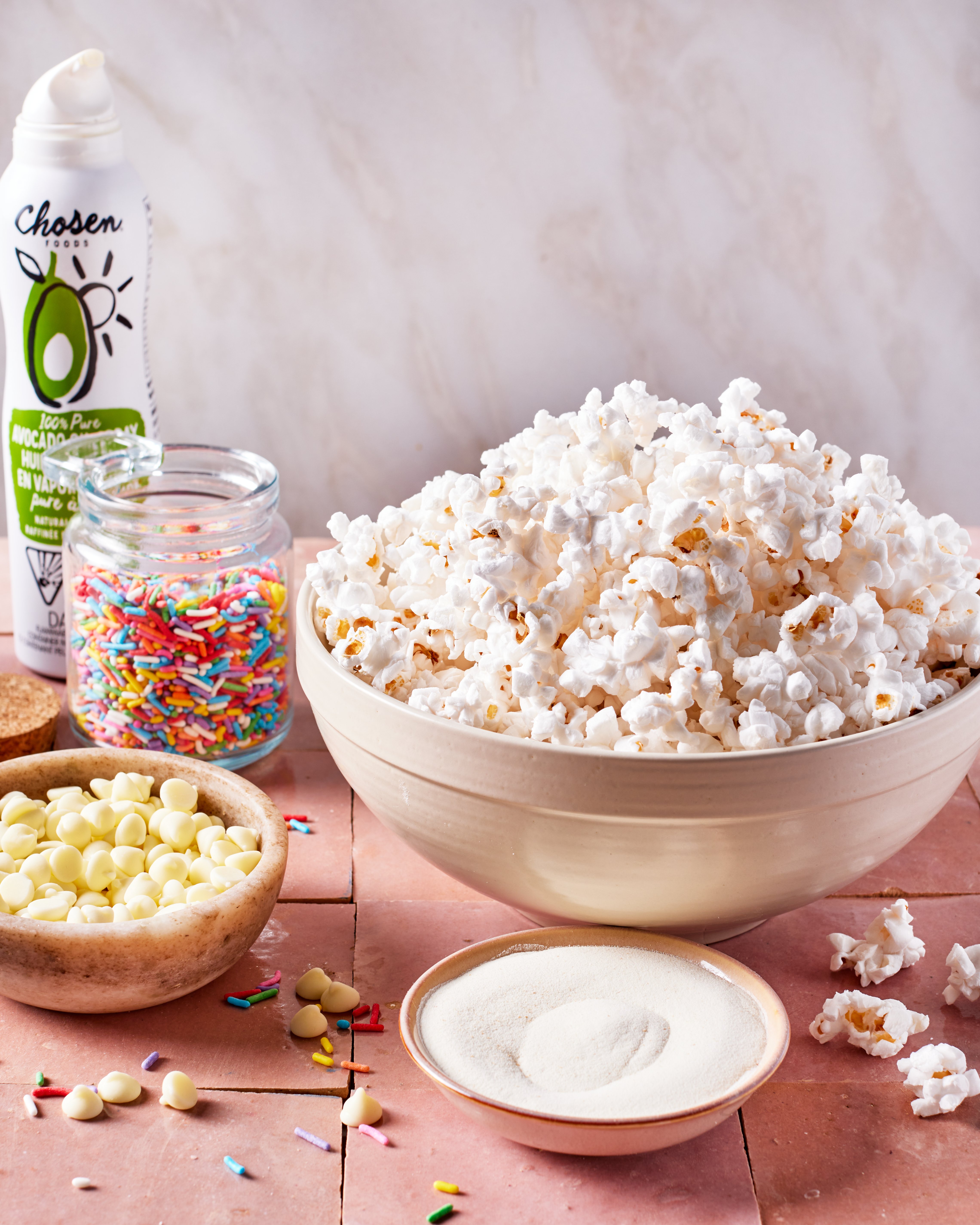 A large bowl of popcorn is surrounded by a small bowl of white chocolate chips, a jar of colorful sprinkles, a dish of white powder, and a can of avocado oil spray on a pink tile surface.