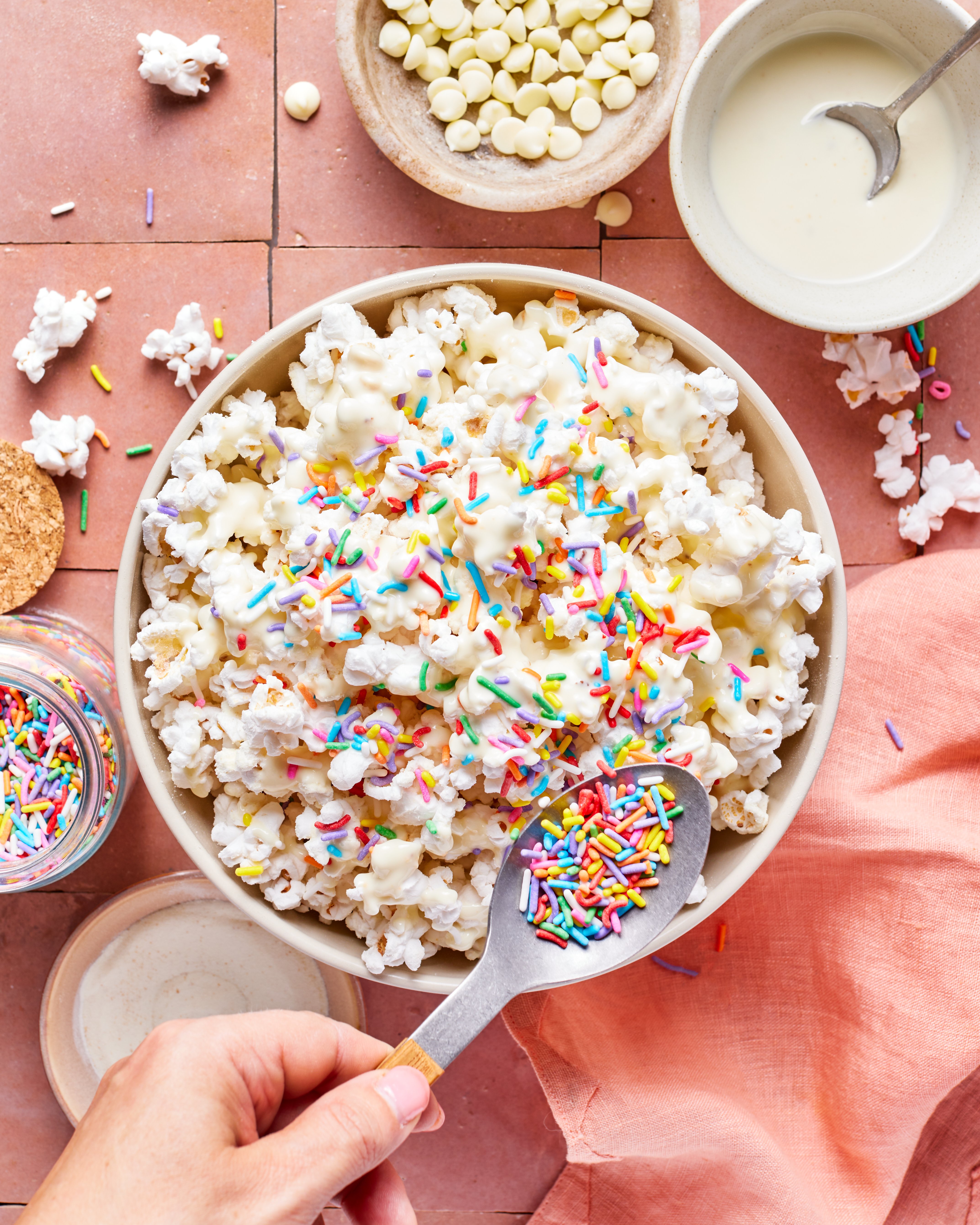 A bowl of popcorn topped with white chocolate and colorful sprinkles sits on a pink surface. A hand adds more sprinkles with a spoon. Nearby are a small bowl of white chocolate chips and a jar of sprinkles.