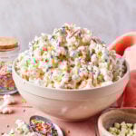 A bowl of popcorn topped with colorful sprinkles sits on a pink surface. A spoon with more sprinkles, a jar of sprinkles, white chocolate chips, and a peach napkin are nearby.