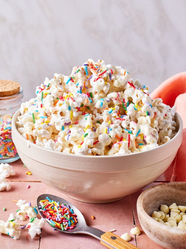 A bowl of popcorn topped with colorful sprinkles sits on a pink surface. A spoon with more sprinkles, a jar of sprinkles, white chocolate chips, and a peach napkin are nearby.