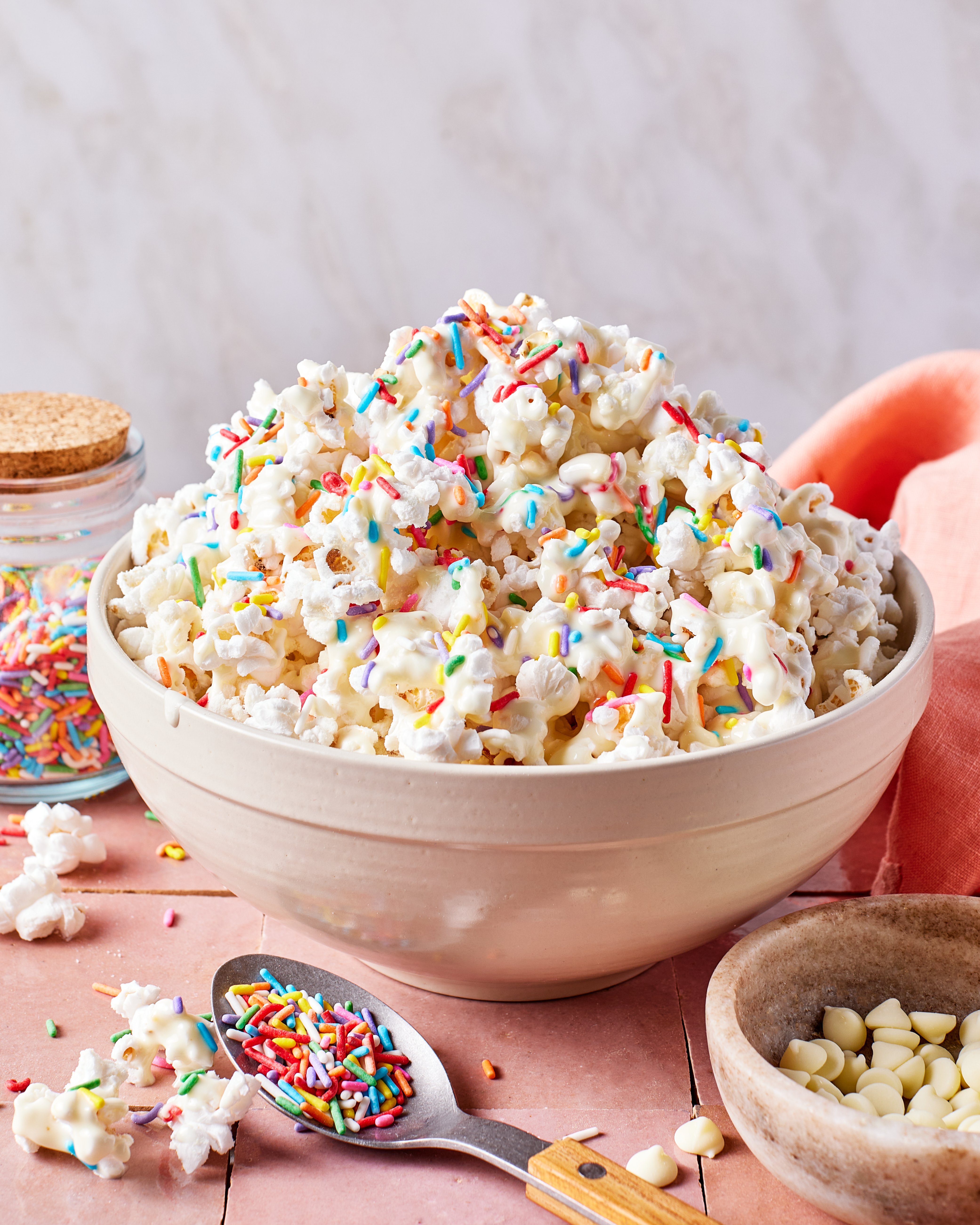 A bowl of popcorn topped with colorful sprinkles sits on a pink surface. A spoon with more sprinkles, a jar of sprinkles, white chocolate chips, and a peach napkin are nearby.