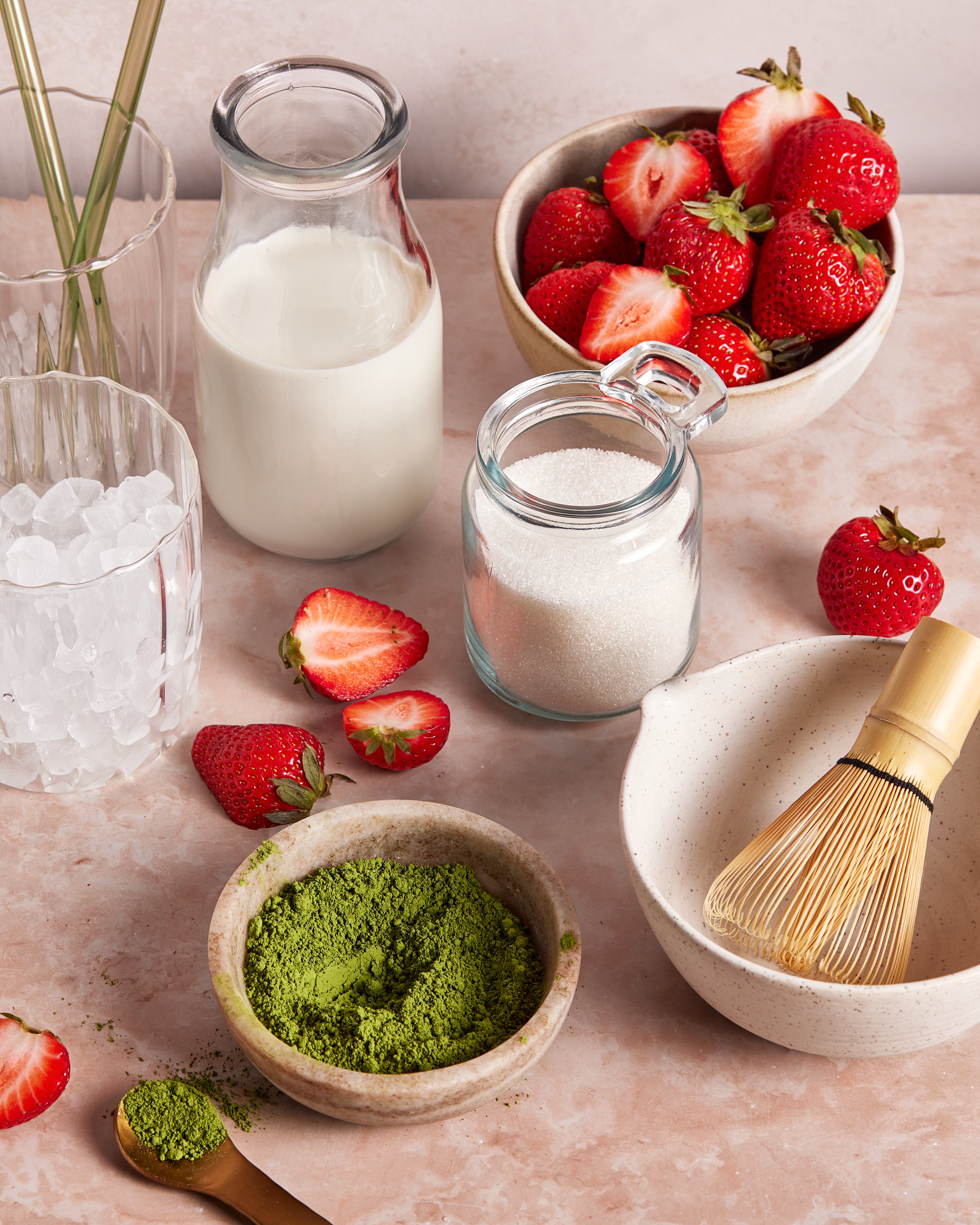 A flat lay of ingredients on a pink surface, including fresh strawberries, a jar of milk, a jar of sugar, a glass with ice, a bowl of matcha powder, a bamboo whisk, and a few sliced strawberries.