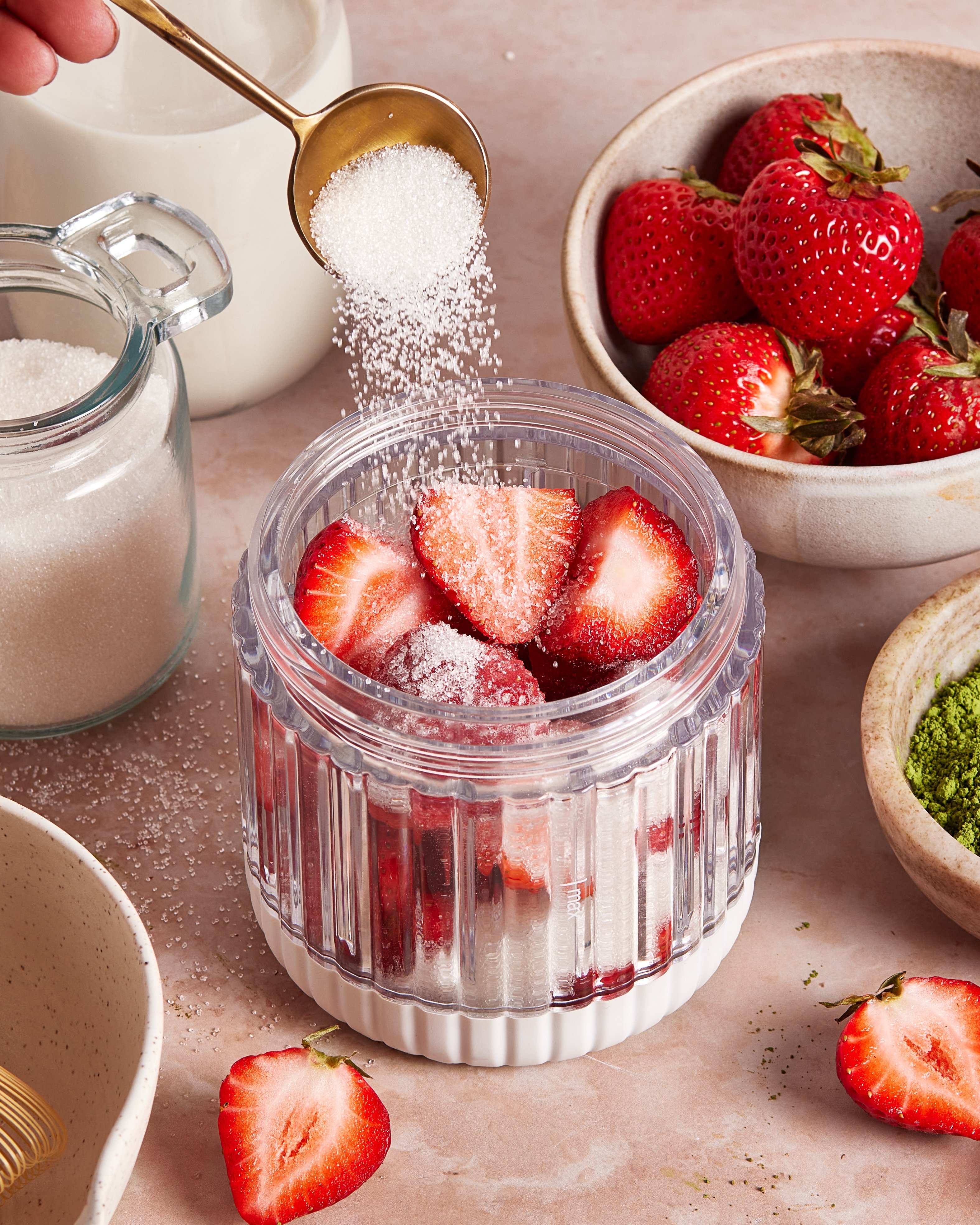 A hand sprinkles sugar from a spoon over sliced strawberries in a glass container, surrounded by bowls of fresh strawberries, sugar, and green matcha powder on a pink countertop.