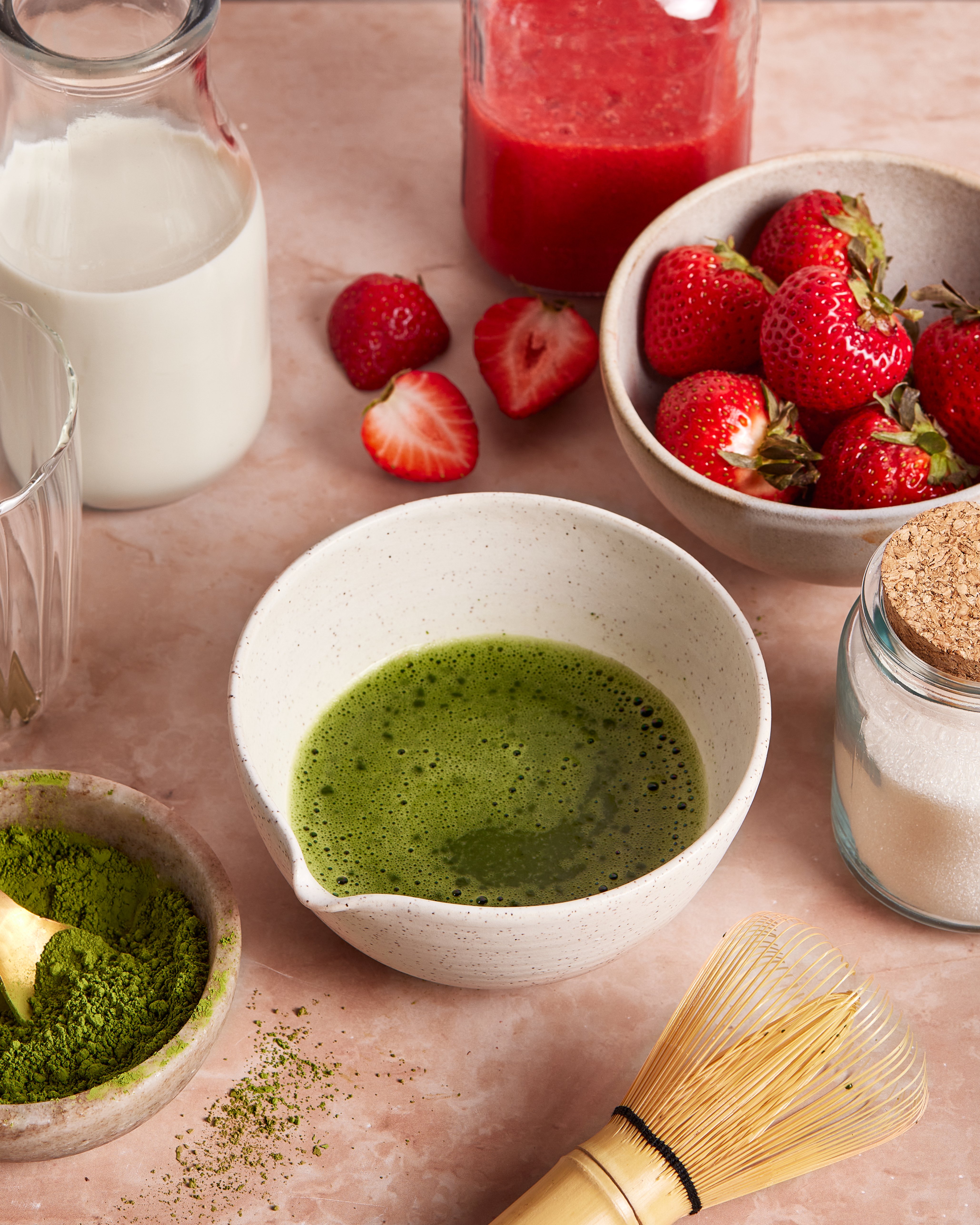 A bowl of whisked matcha with a bamboo whisk, matcha powder, fresh strawberries, a jar of milk, a glass, a jar of sugar, and a bowl of strawberry puree on a light pink surface.