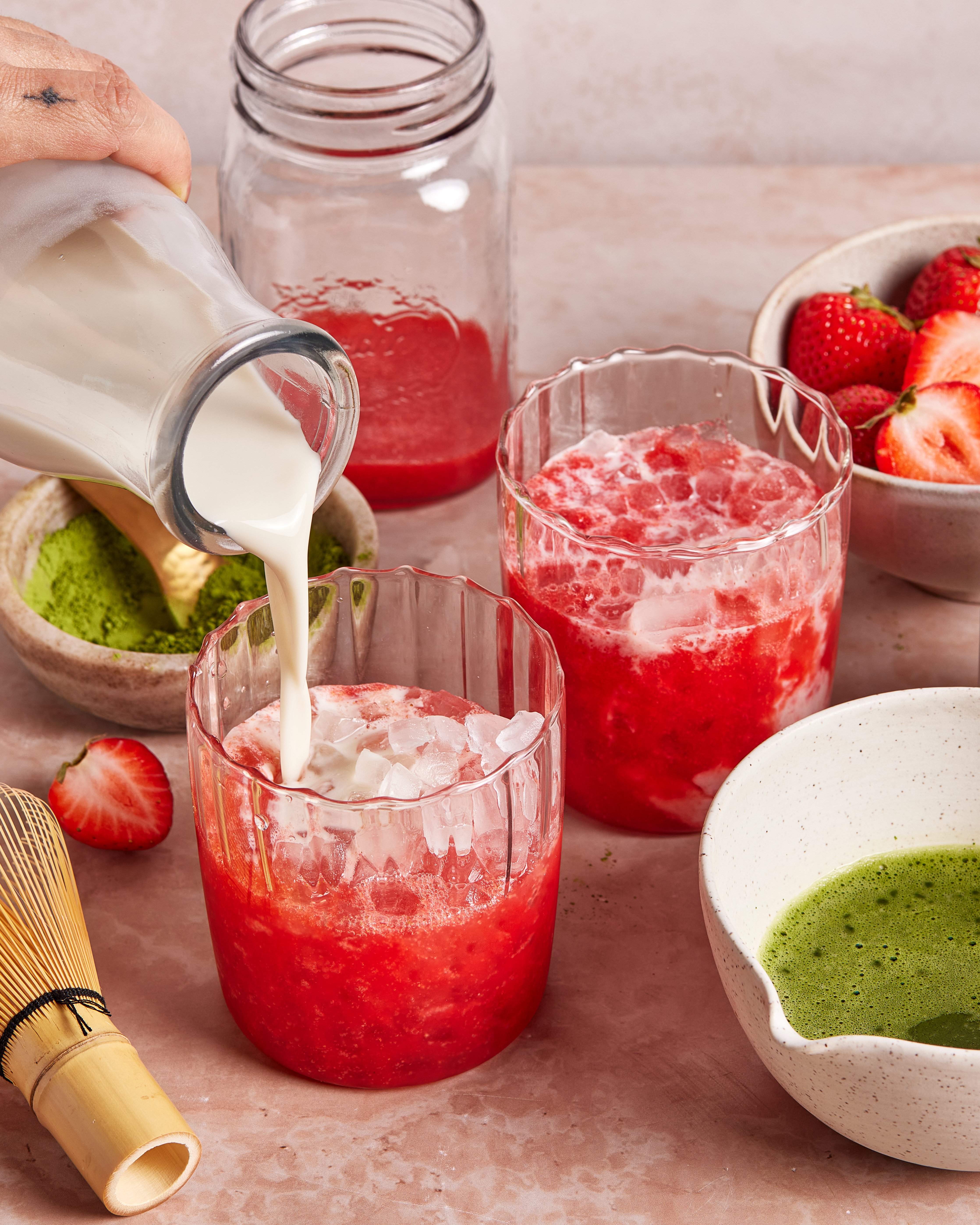 A hand pours milk into a glass of iced strawberry mixture. Next to it is another glass with the same mixture, a bowl of matcha powder, a bowl of fresh strawberries, and a bamboo whisk on a light surface.