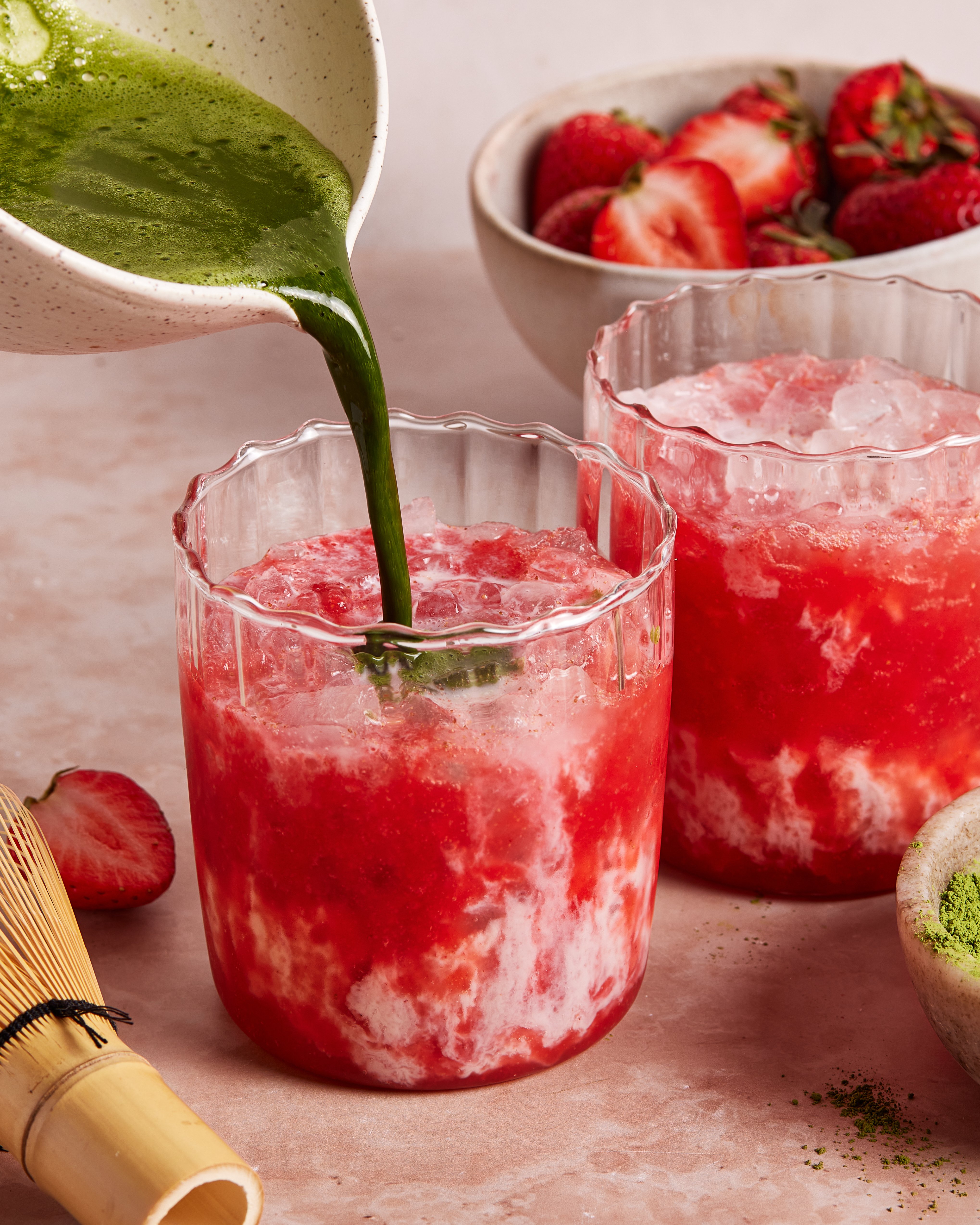 A green matcha mixture is being poured into a glass of iced strawberry drink, with another similar drink, fresh strawberries, a bamboo whisk, and a bowl of matcha powder nearby on a pink surface.