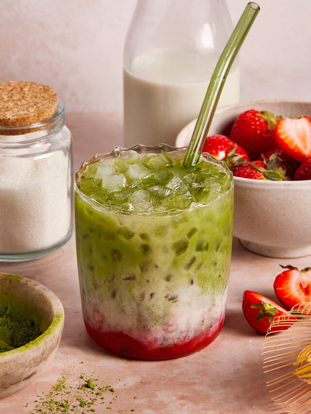 A glass of iced matcha latte with layers of green matcha, milk, and red strawberry puree sits on a table with a glass straw. Surrounding it are bowls of sugar, strawberries, a bottle of milk, and a dish of matcha powder.