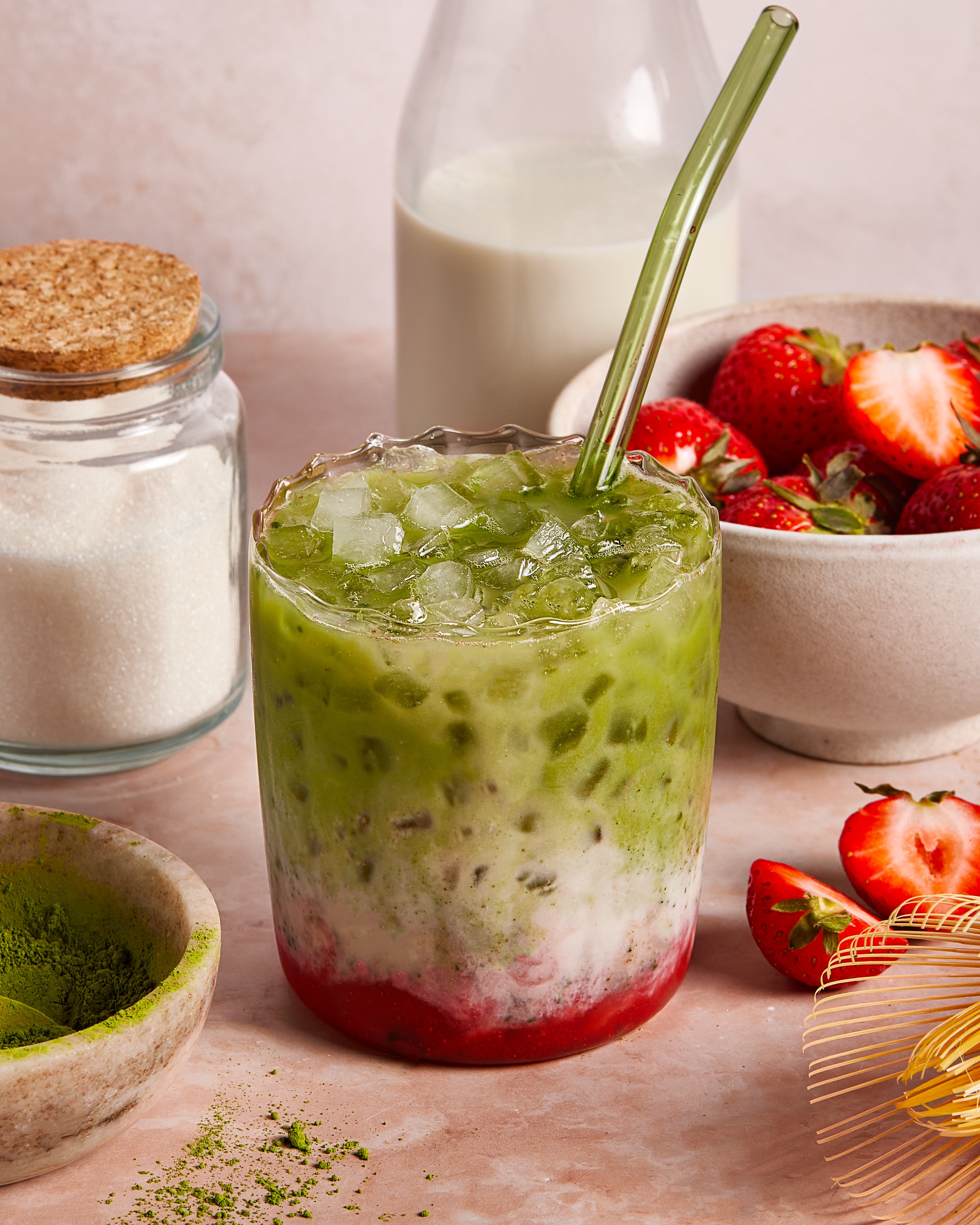 A glass of iced matcha latte with layers of green matcha, milk, and red strawberry puree sits on a table with a glass straw. Surrounding it are bowls of sugar, strawberries, a bottle of milk, and a dish of matcha powder.