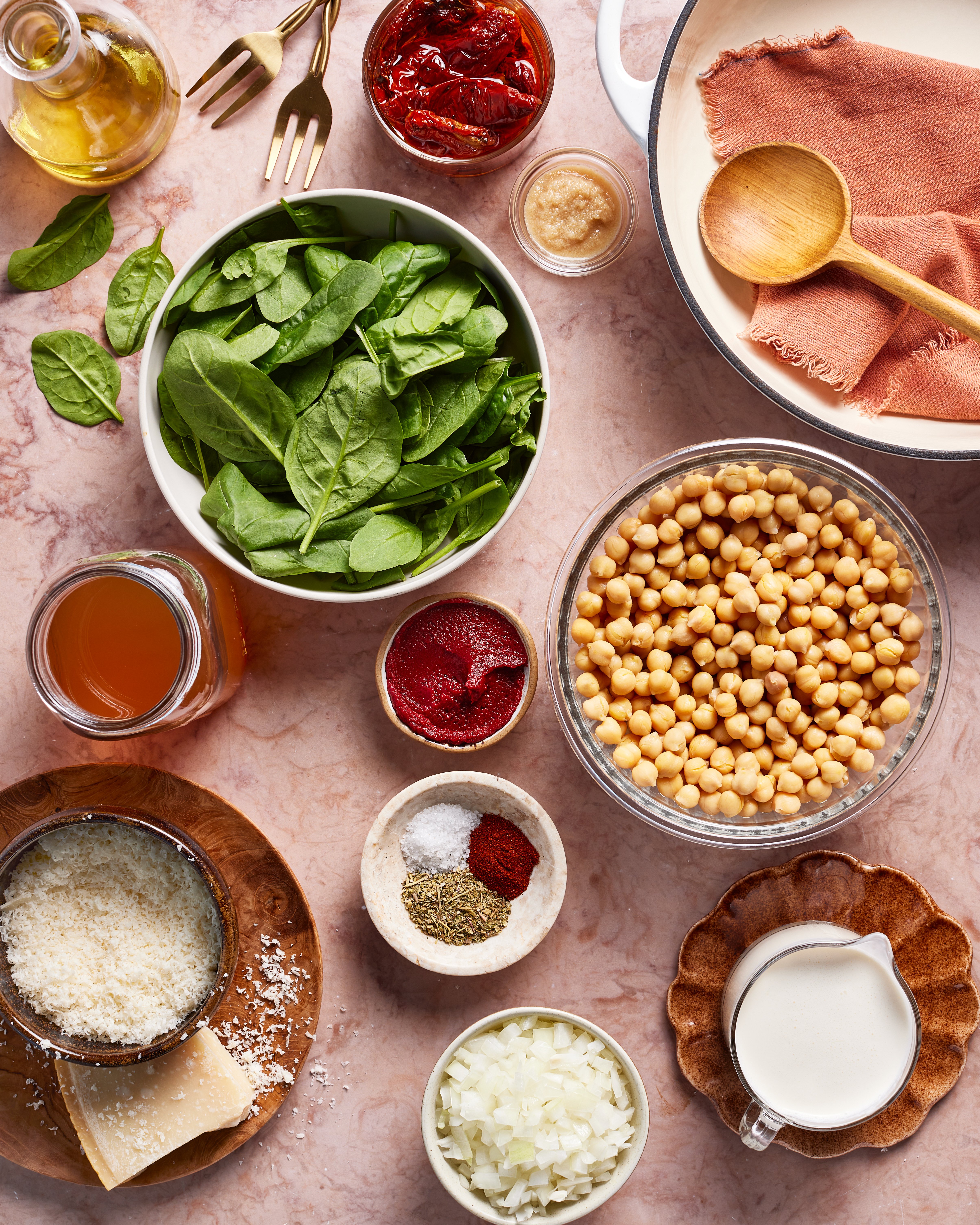 An overhead view of various ingredients on a pink surface, including spinach, chickpeas, chopped onion, grated cheese, spices, tomato paste, broth, plant-based cream, olive oil, sun-dried tomatoes, and a white pot with a wooden spoon.