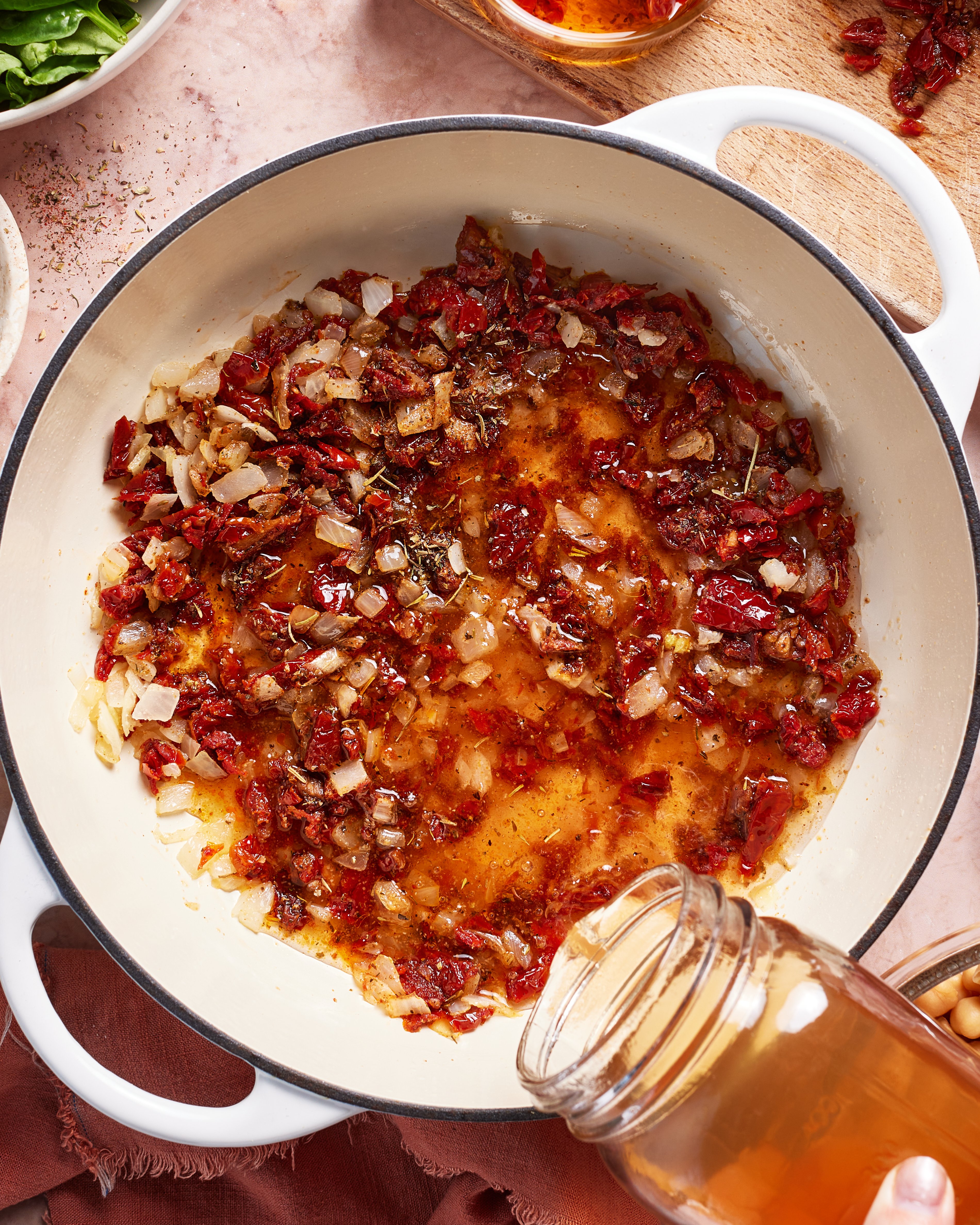 A white pot contains sautéed onions, sun-dried tomatoes, and herbs, with broth being poured in from a glass jar. The scene suggests the start of a cooking process, with additional ingredients visible nearby.