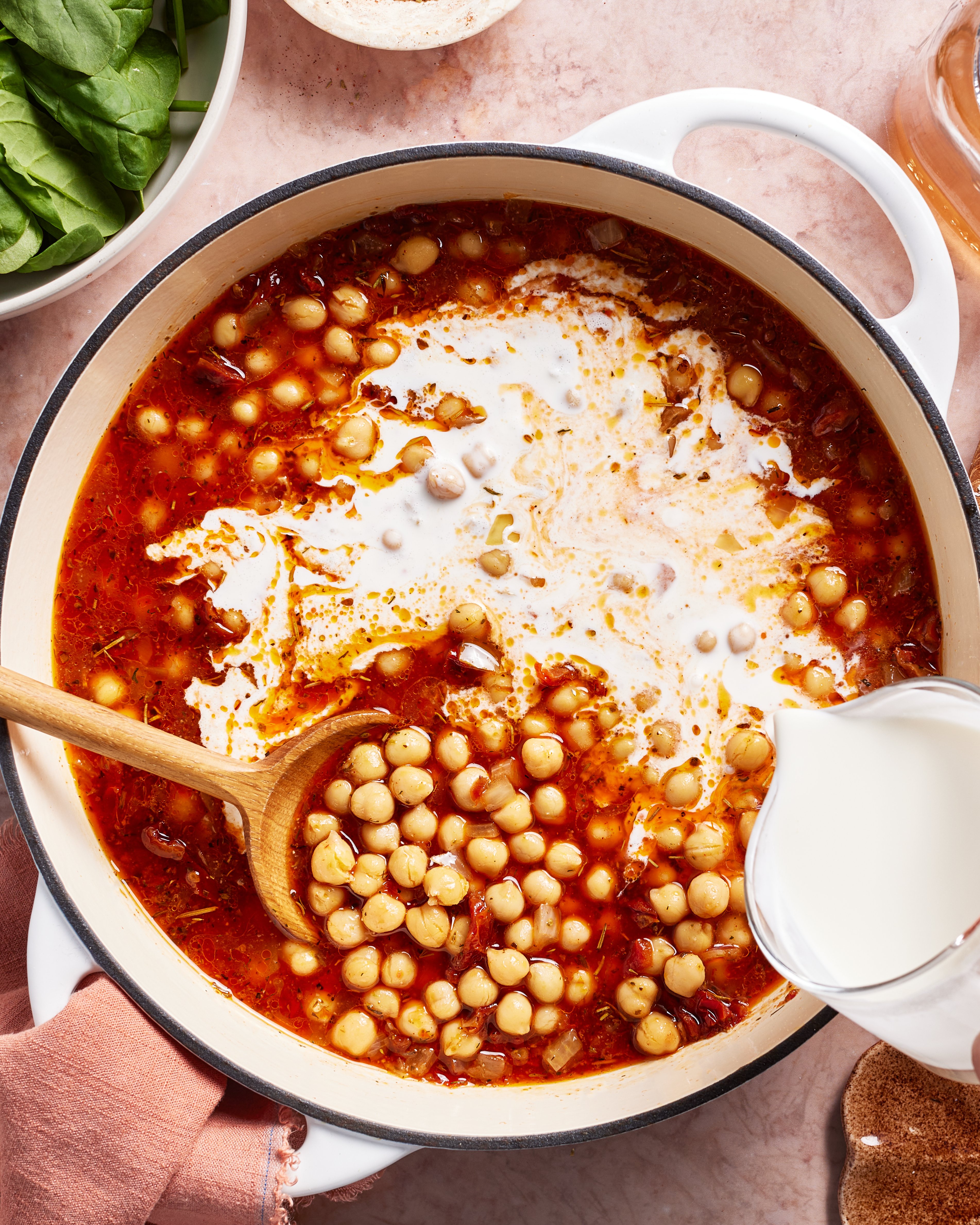 A pot of chickpea stew with a tomato-based broth and a swirl of coconut milk being added, stirred with a wooden spoon. Fresh spinach and other ingredients are visible nearby on a light countertop.