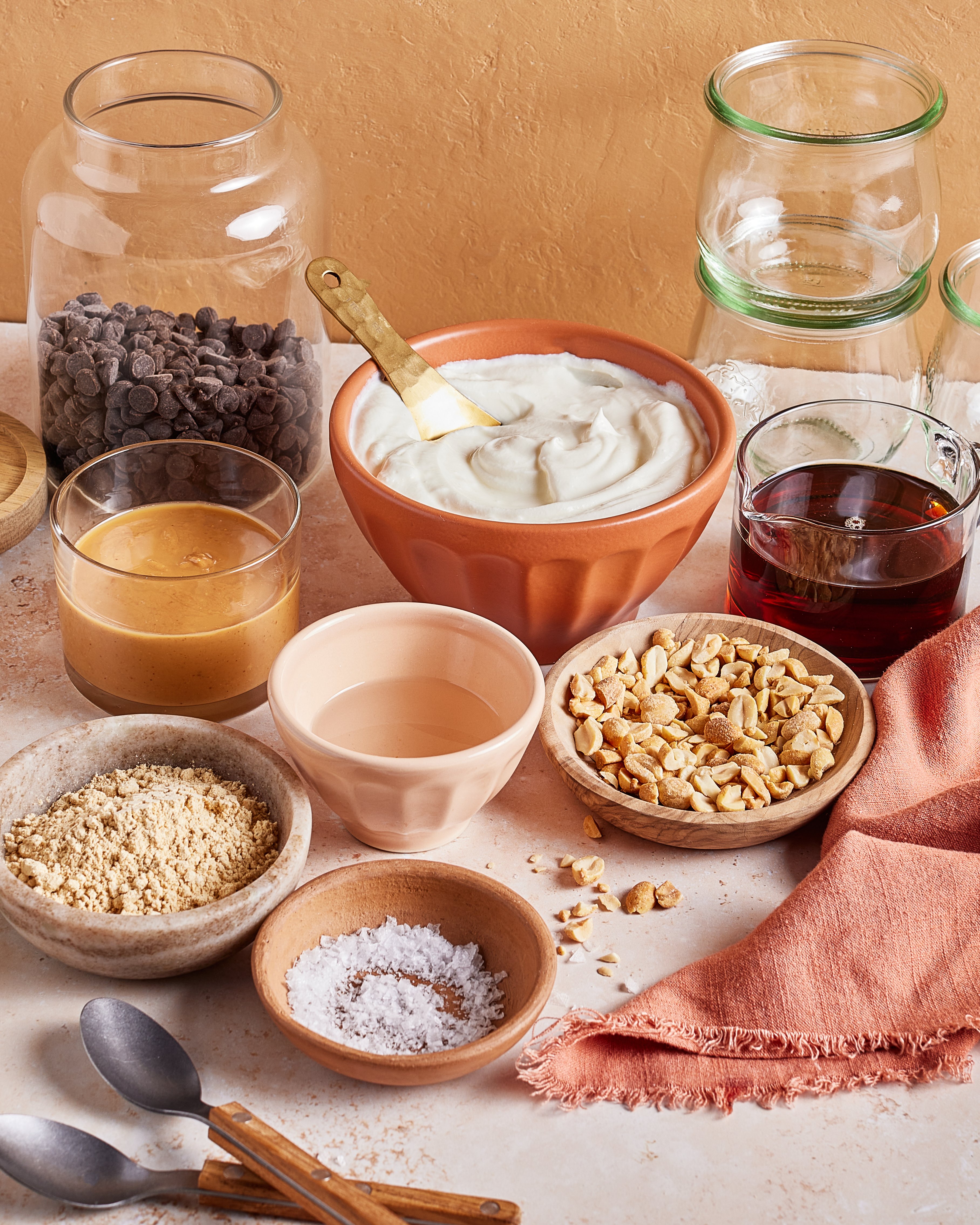 A tabletop displays ingredients in bowls and jars, including yogurt, chopped nuts, chocolate chips, peanut butter, a clear liquid, brown liquid, a powder, and flaked salt, with spoons and an orange cloth nearby.