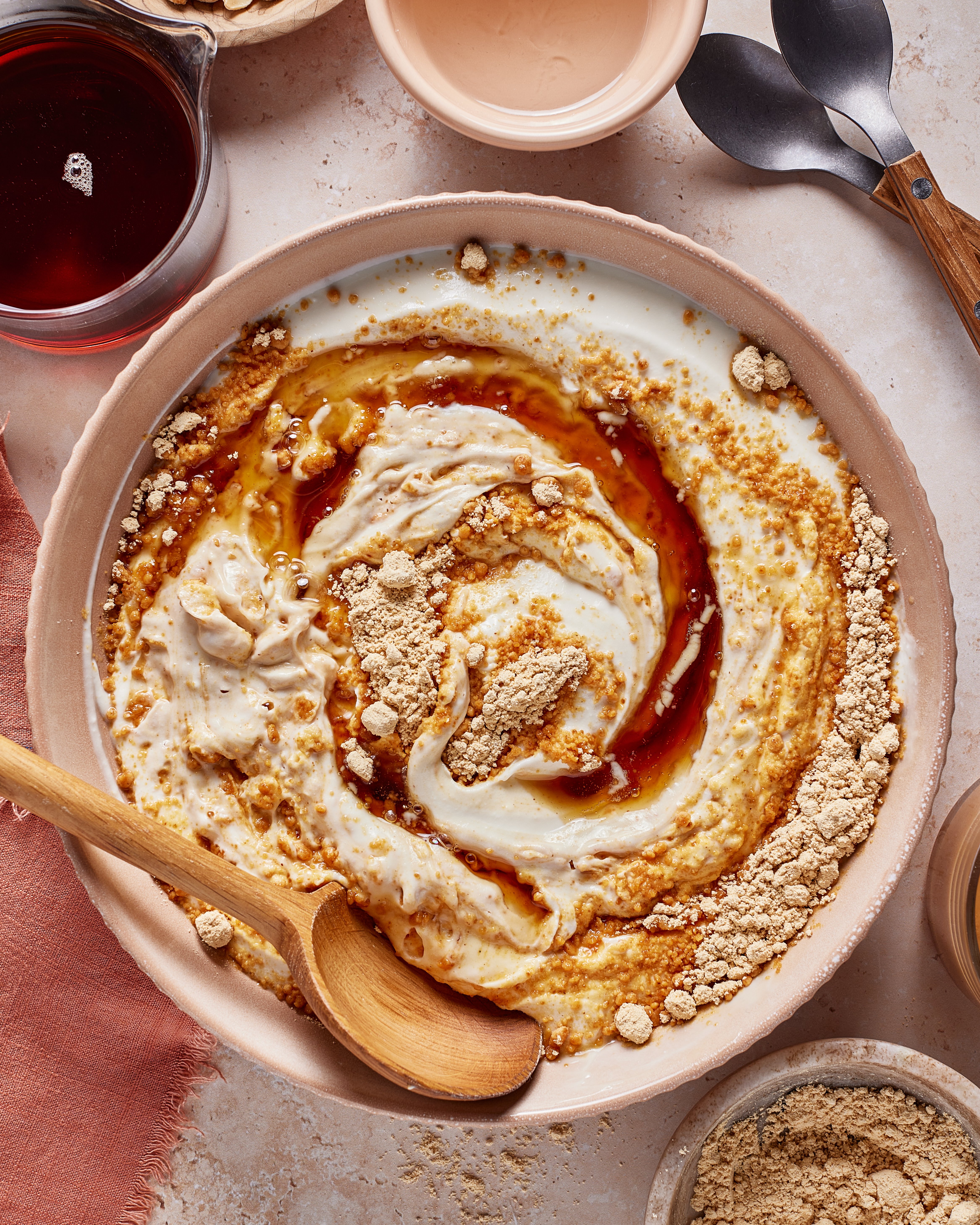 A bowl of yogurt swirled with brown syrup and sprinkled with beige powder, accompanied by a wooden spoon. Nearby are small bowls with extra syrup and powder, and two metal spoons on the side.