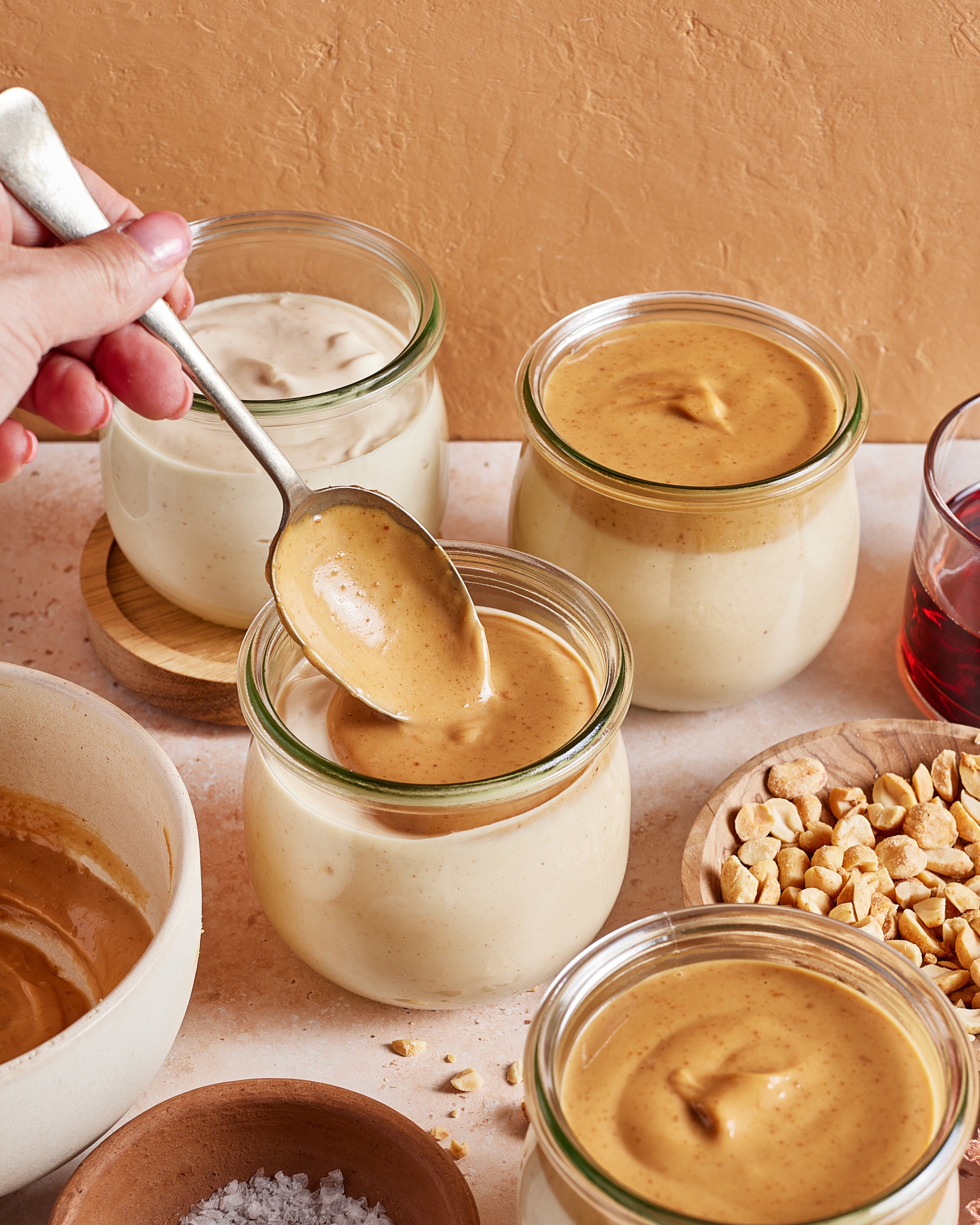 A hand holds a spoon above a jar of creamy peanut butter, surrounded by jars of peanut butter, a bowl of peanuts, a bowl of salt, and a glass of red liquid on a light surface.