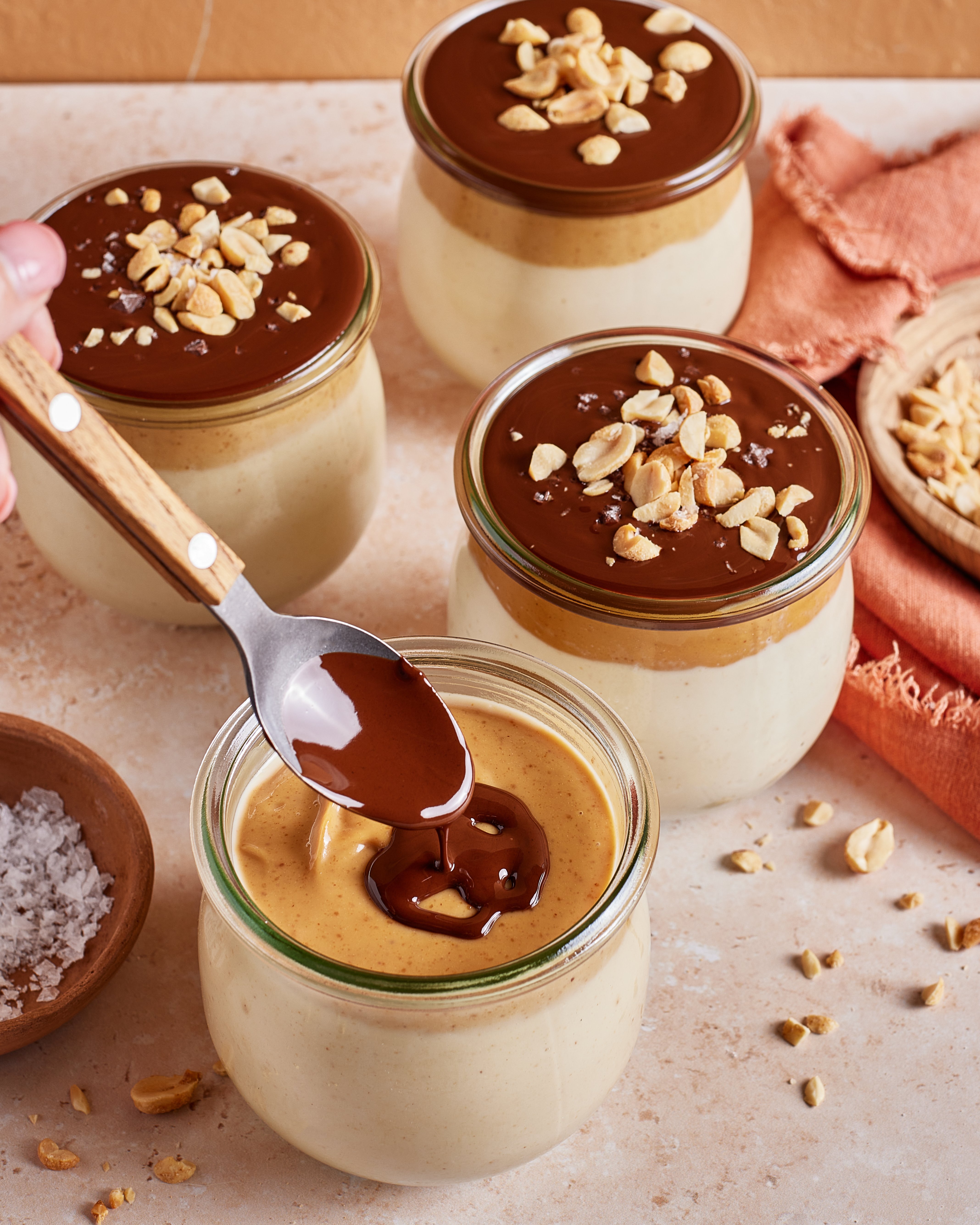 Four jars of creamy dessert, each topped with chocolate and chopped peanuts. A hand holds a spoon drizzling chocolate over one jar. Peanuts, salt, and an orange cloth are nearby on a light surface.