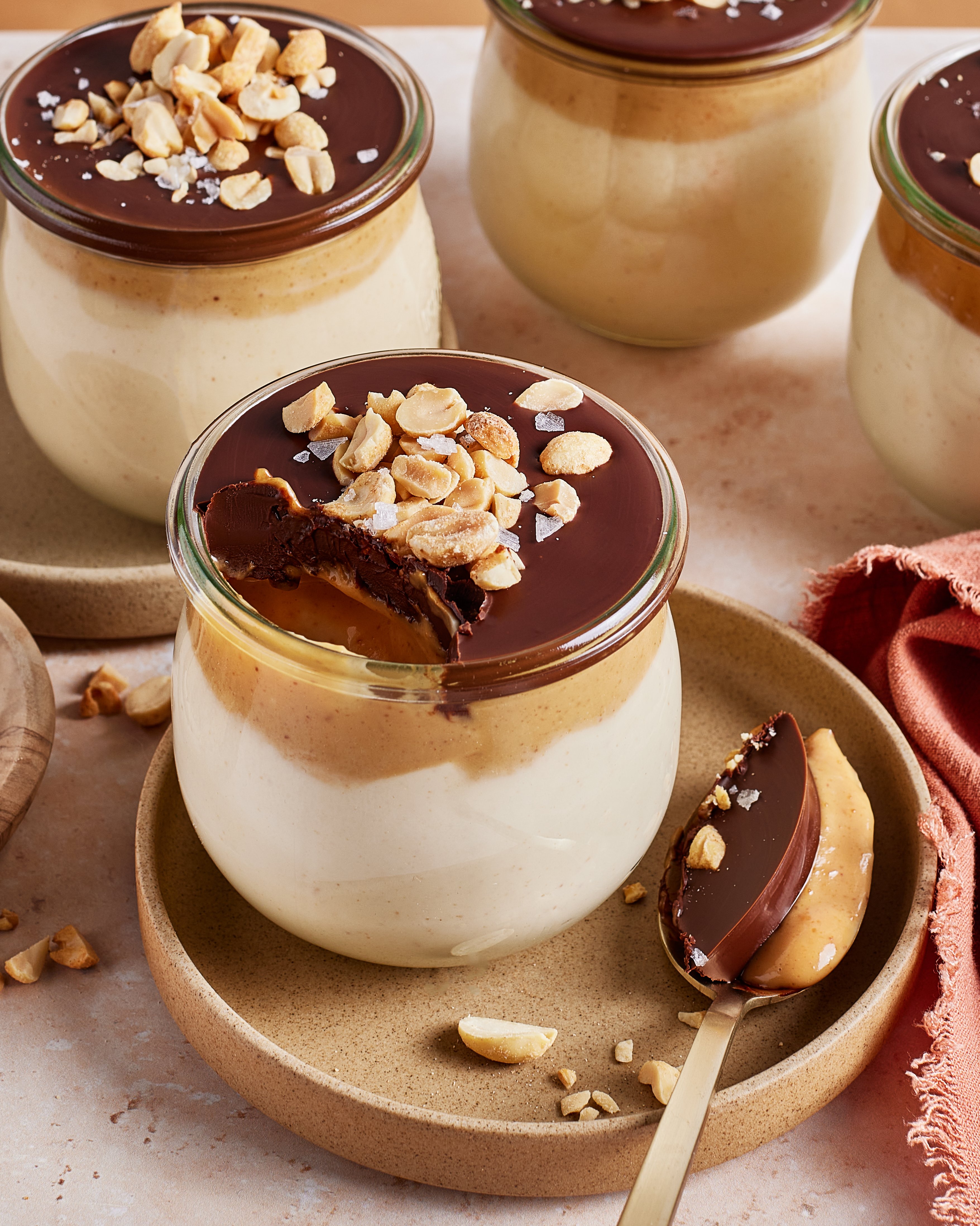 Four glass jars filled with creamy mousse, topped with a layer of chocolate, peanut butter, and chopped peanuts. One jar has a spoonful taken out, showing the dessert layers on a beige plate.