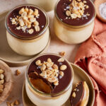 Four glass jars of creamy dessert topped with a chocolate layer, salted peanuts, and flaky salt. One jar has a spoonful taken out, showing the smooth interior. A spoon with dessert and a napkin are nearby.