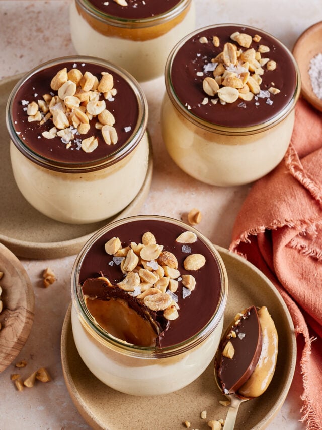 Four glass jars of creamy dessert topped with a chocolate layer, salted peanuts, and flaky salt. One jar has a spoonful taken out, showing the smooth interior. A spoon with dessert and a napkin are nearby.