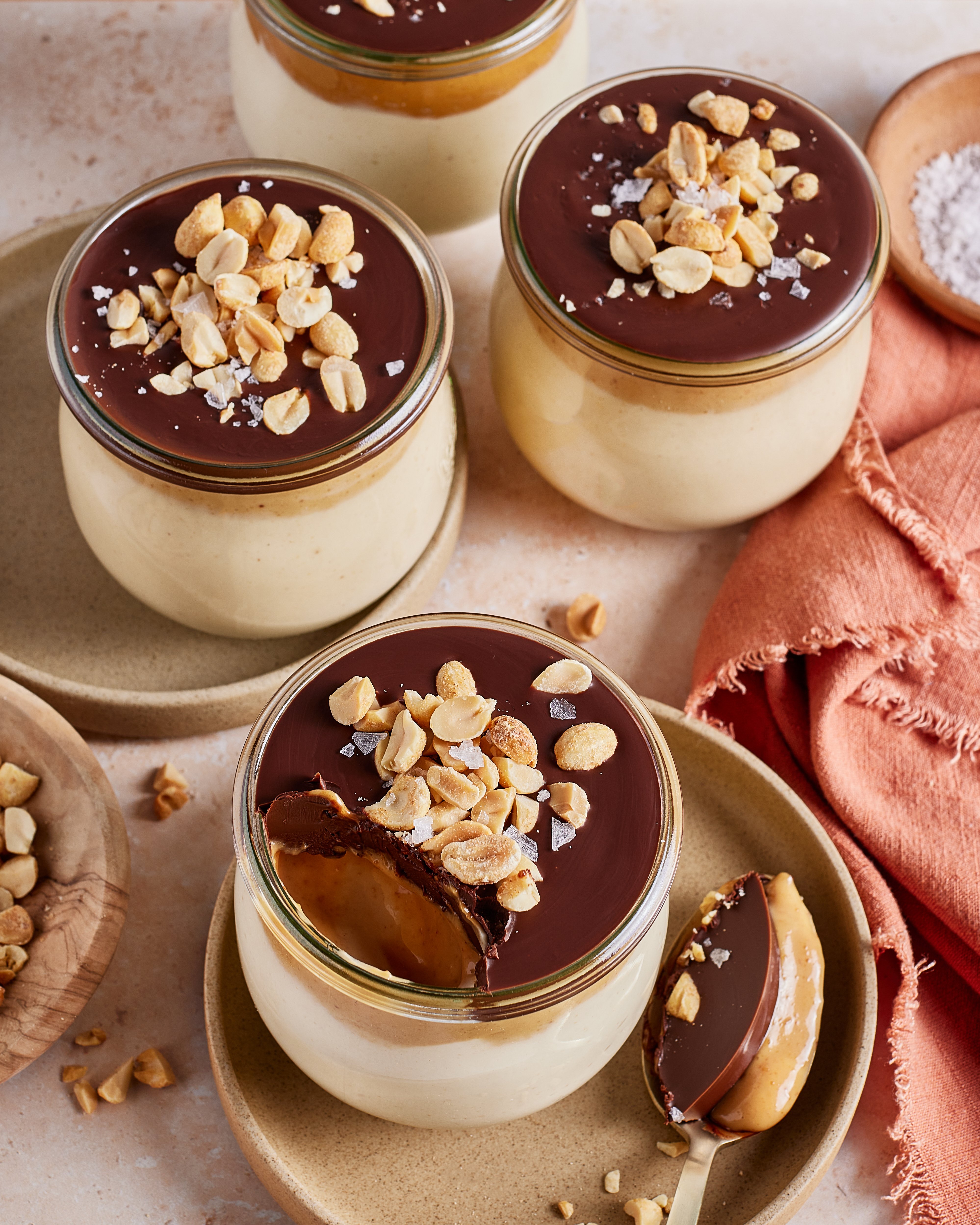 Four glass jars of creamy dessert topped with a chocolate layer, salted peanuts, and flaky salt. One jar has a spoonful taken out, showing the smooth interior. A spoon with dessert and a napkin are nearby.