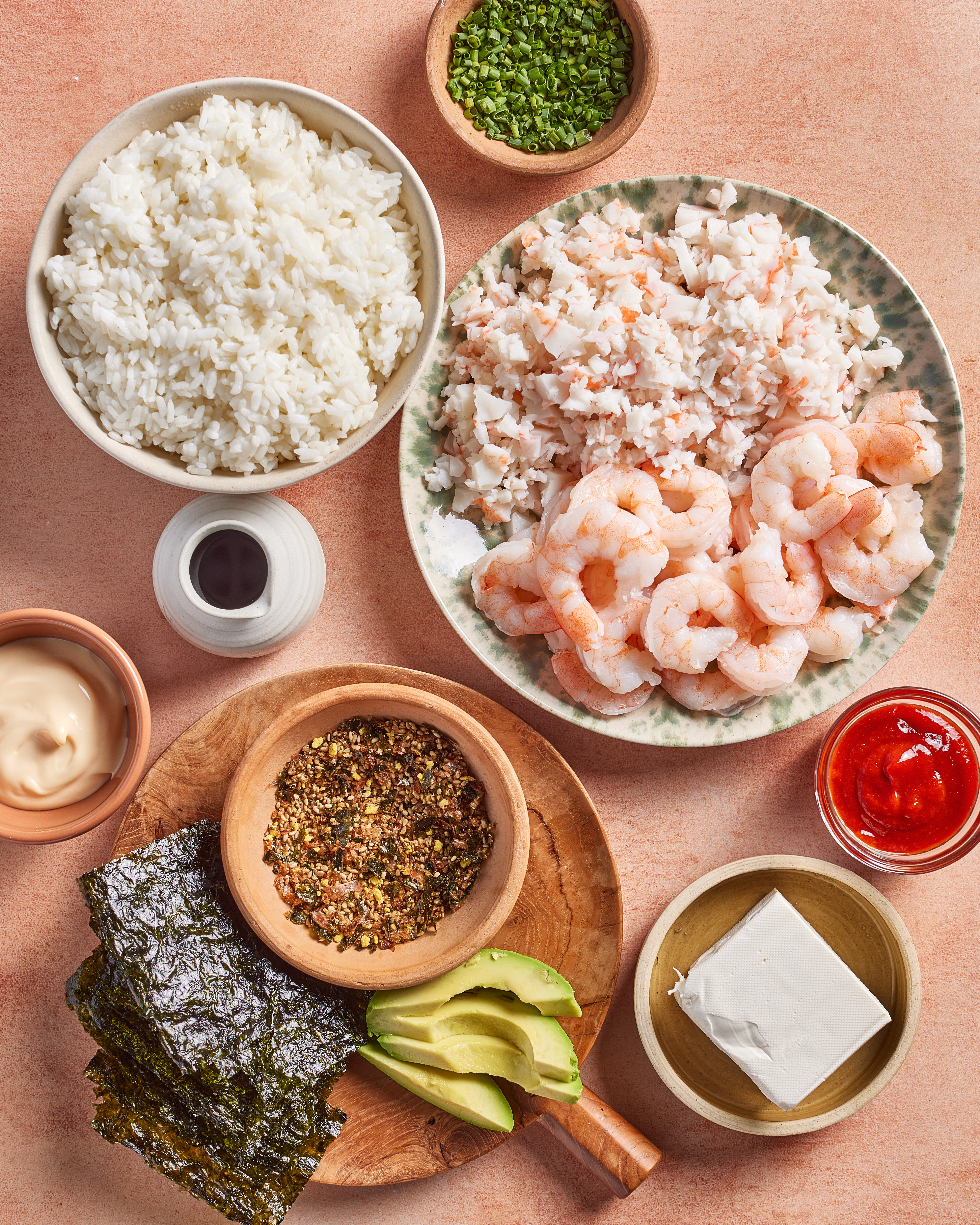 An overhead view of ingredients for sushi: a plate with chopped crab and shrimp, a bowl of rice, bowls of chopped chives, soy sauce, mayonnaise, furikake, sliced avocado, sriracha, nori sheets, and a block of tofu.