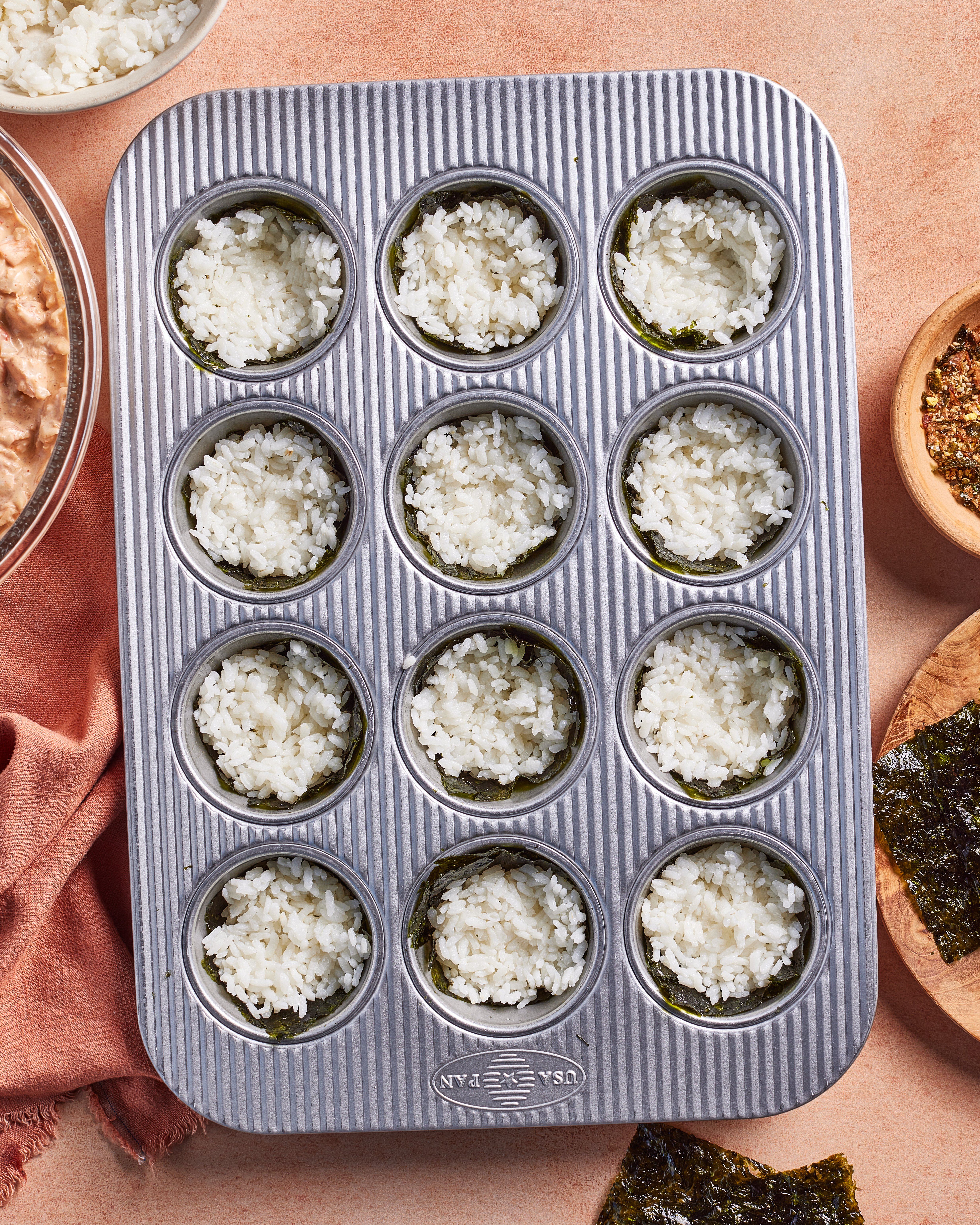 A metal muffin tin with 12 wells, each lined with a sheet of seaweed and filled with white rice. Surrounding the tin are a pink cloth, bowls, and extra seaweed sheets on a beige surface.