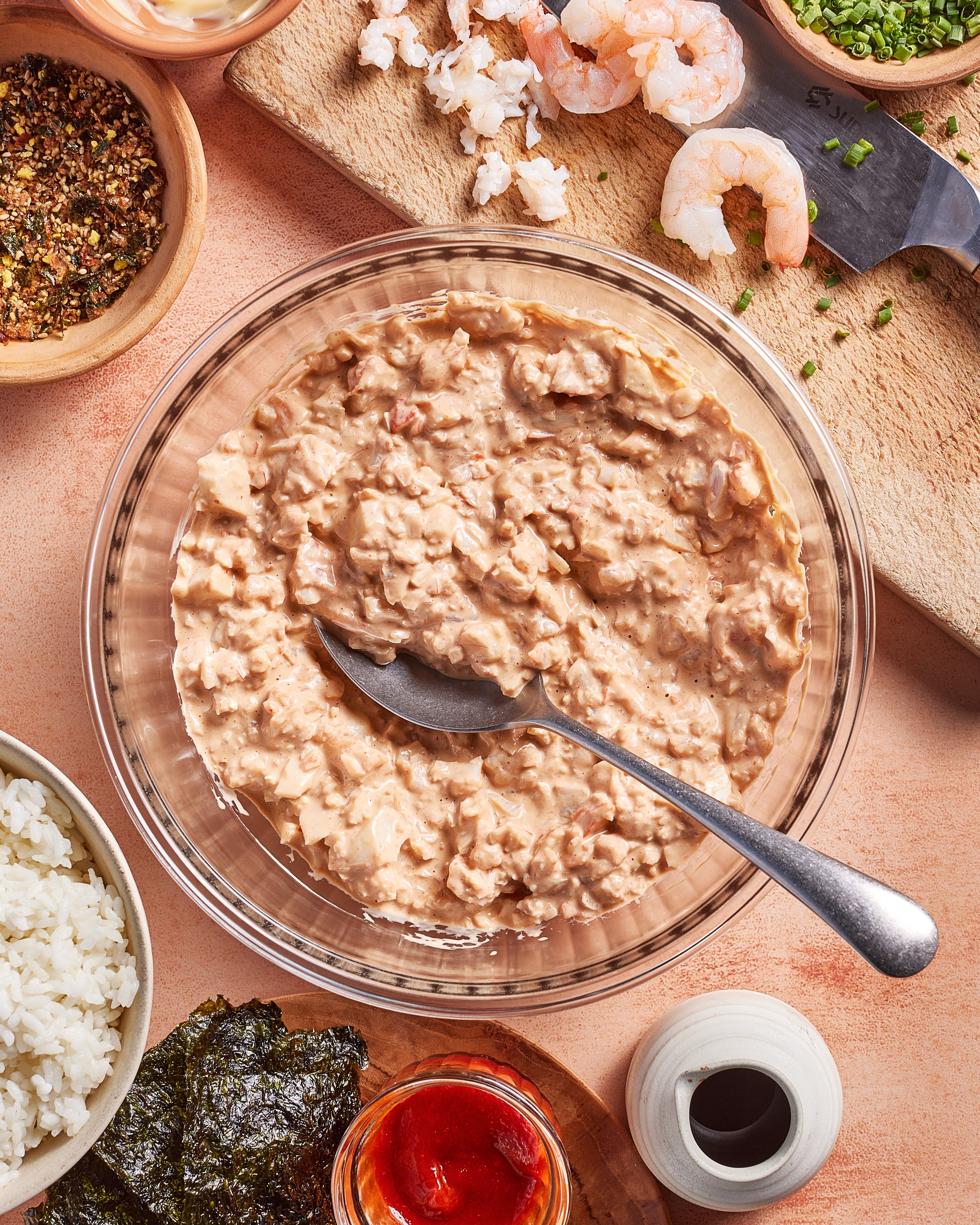 A bowl of creamy shrimp mixture with a spoon in it, surrounded by ingredients like shrimp, chopped herbs, white rice, nori seaweed, spices, and sauce on a light-colored surface.