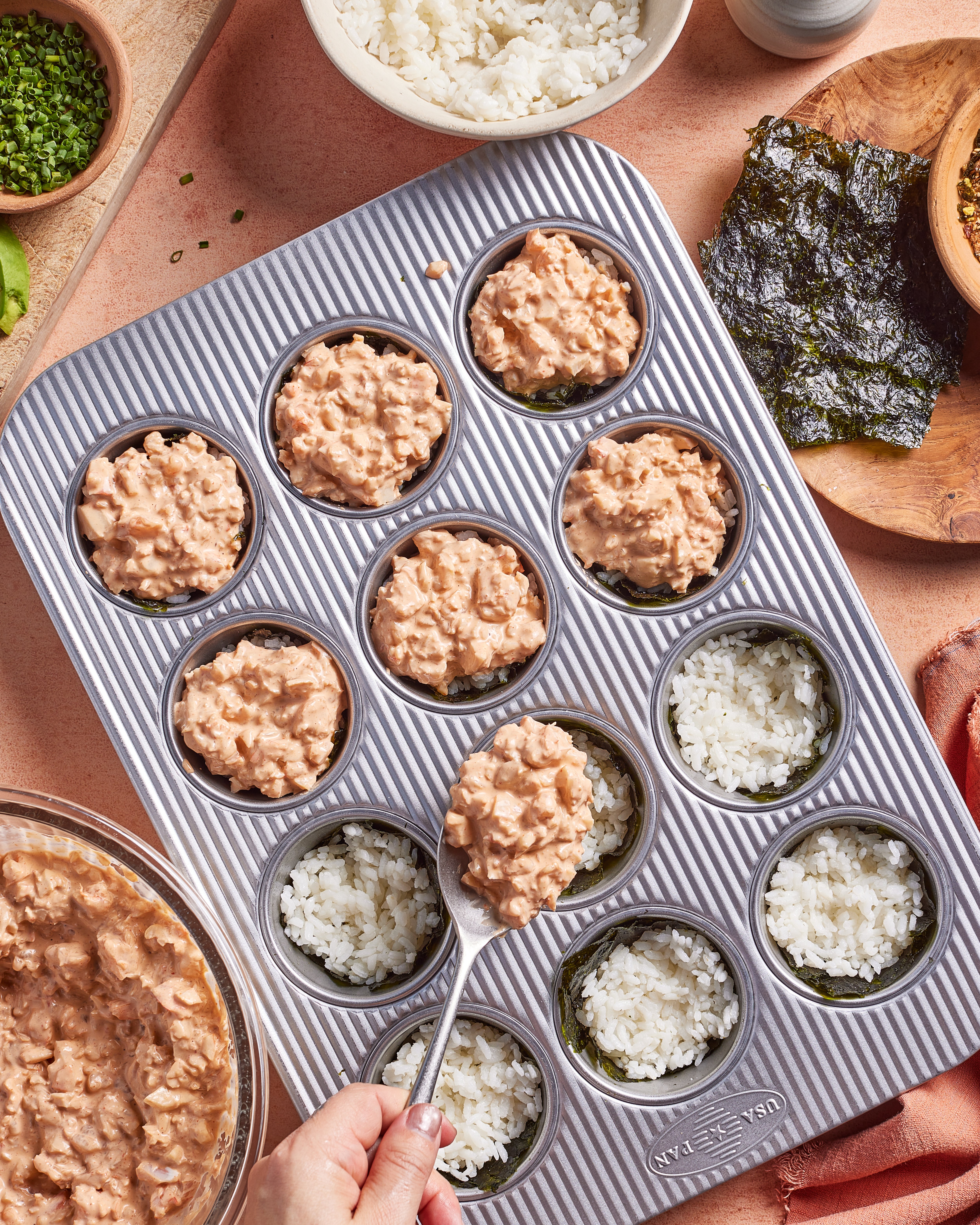 A hand fills a muffin tin lined with seaweed and rice with a tuna mixture. Several cups are already filled, while others contain only rice and seaweed. Bowls of rice and seaweed are nearby.