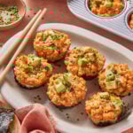 A plate of six Spicy California Roll Baked Sushi Cups topped with avocado and chopped chives, surrounded by chopsticks, a bowl of dipping sauce, a baking tray with more sushi cups, and a small dish of chives.