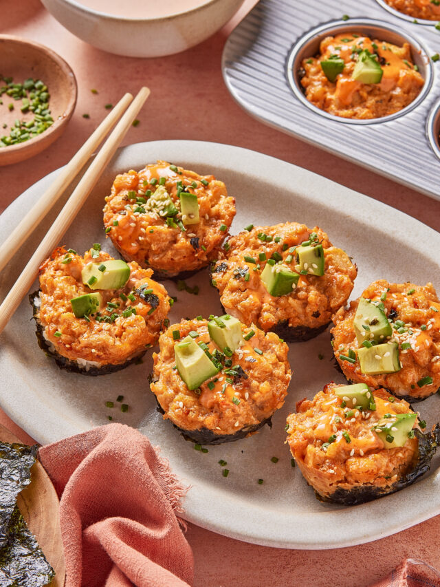 A plate of six Spicy California Roll Baked Sushi Cups topped with avocado and chopped chives, surrounded by chopsticks, a bowl of dipping sauce, a baking tray with more sushi cups, and a small dish of chives.