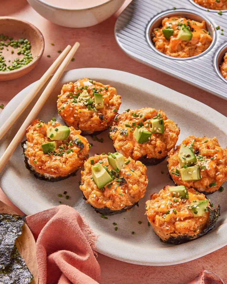 A plate of six Spicy California Roll Baked Sushi Cups topped with avocado and chopped chives, surrounded by chopsticks, a bowl of dipping sauce, a baking tray with more sushi cups, and a small dish of chives.