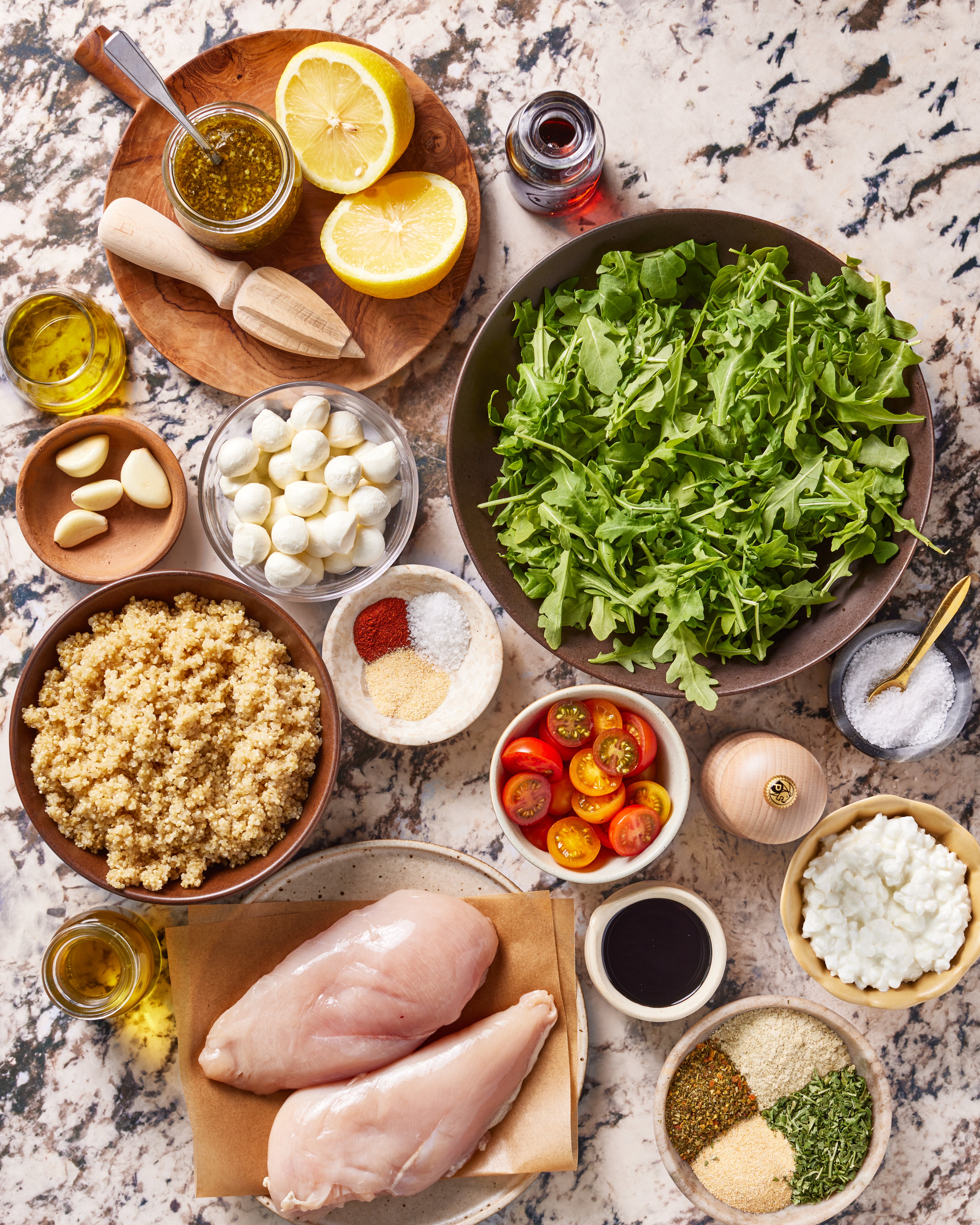 A top-down view of ingredients on a marble counter including raw chicken breasts, arugula, quinoa, cherry tomatoes, mozzarella balls, lemon halves, garlic cloves, spices, cottage cheese, and small bowls of sauces and oils. (Copycat Sweetgreen Chicken Caprese Salad)